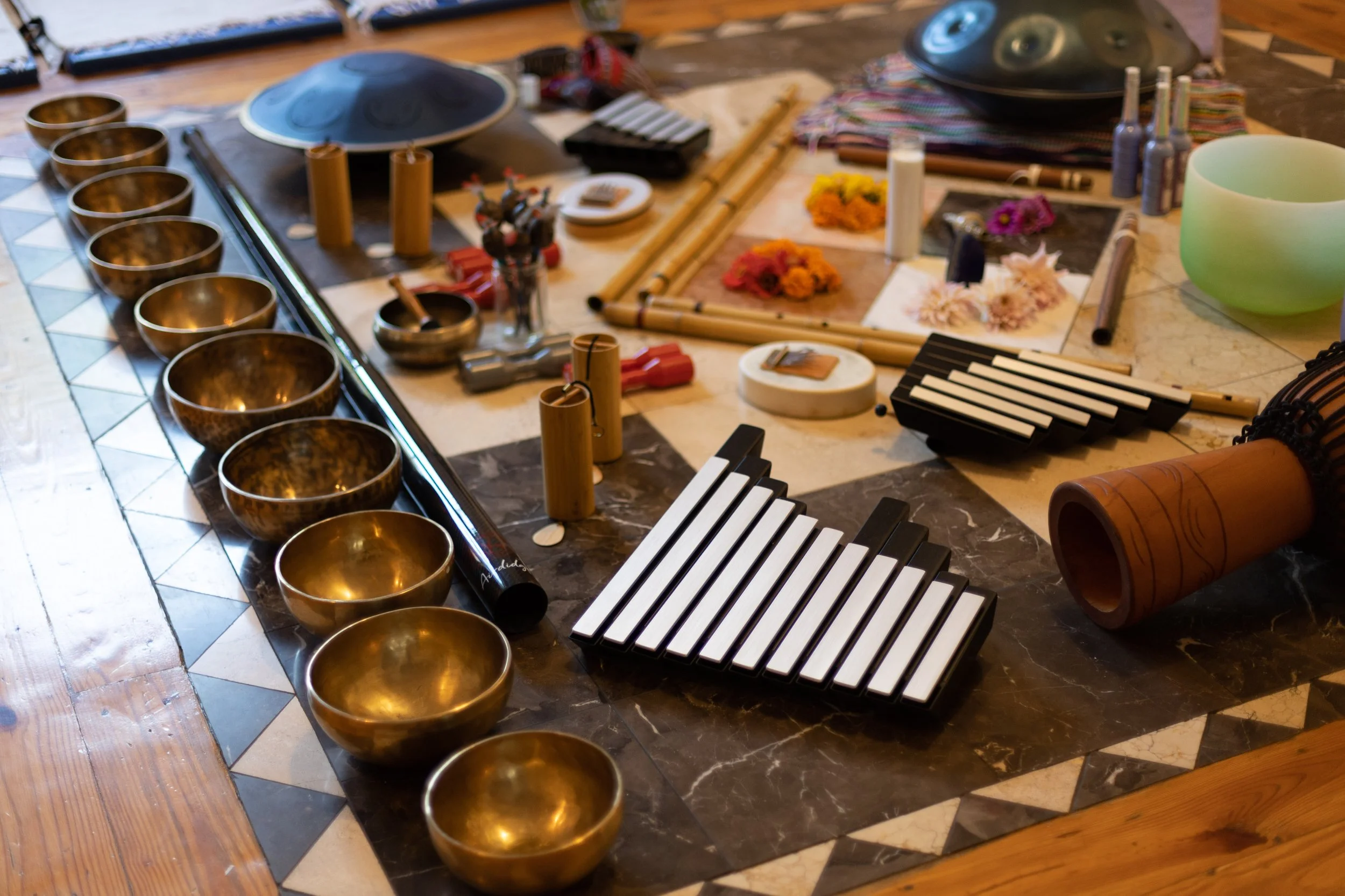 Various musical instruments including singing bowls, a xylophone, kalimbas, and a handpan are arranged on a table with a tile and wooden floor.