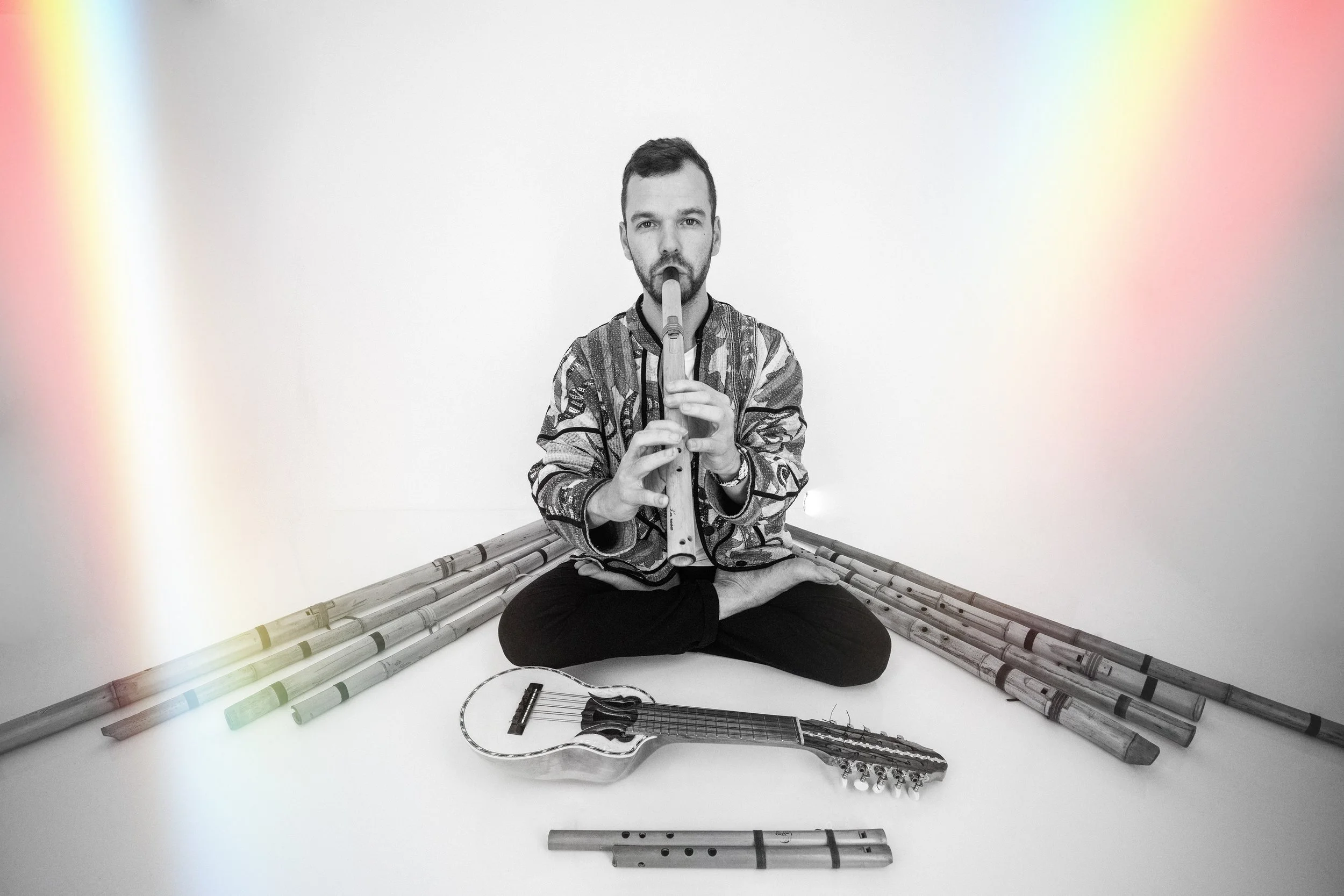 Adrian DiMatteo cross-legged on the floor playing a native flute. Surrounding him are bamboo flutes from Peru of different sizes and a small guitar, with a white background.