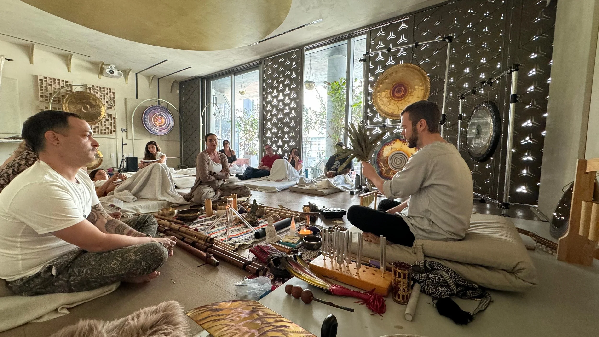 A meditation or sound healing session with a group of people in a serene indoor space. Participants are seated or lying down, surrounded by musical instruments and spiritual objects, while a person at the front appears to lead the session. Large gongs and decorative elements adorn the background.