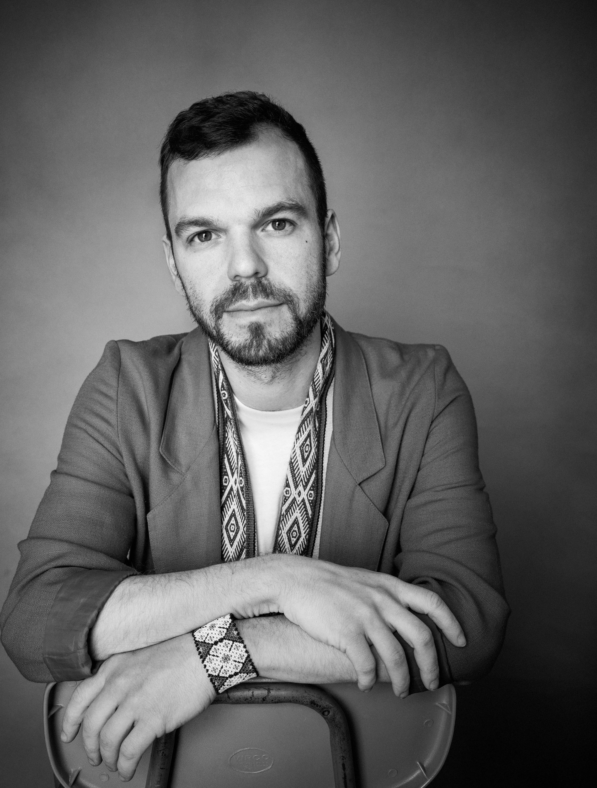 A black and white portrait of a man with short dark hair, beard, and mustache. He's wearing a jacket over a shirt with a patterned scarf around his neck. He is seated with his arms crossed on the table in front of him, and the background is plain and slightly gradient.