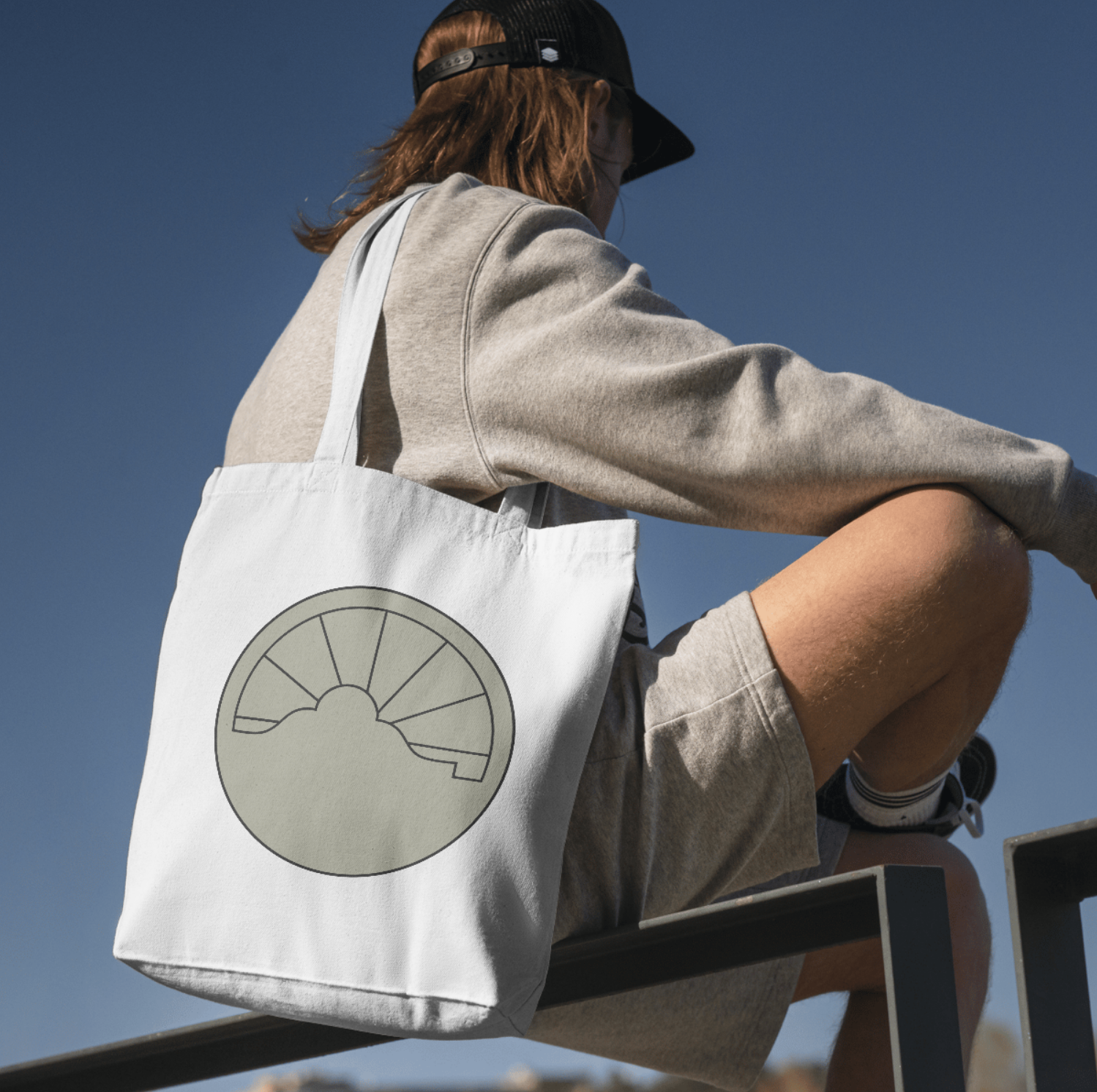 A person wearing a black cap, gray sweatshirt, and shorts with striped socks, sitting on a ledge outdoors against a clear blue sky, carrying a white tote bag with a minimalist design of a sun and cloud.