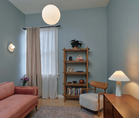 Living room with blue walls, pink sofa, wooden bookshelf with decorations, gray armchair, wooden side table with white lamp, and cream curtains.
