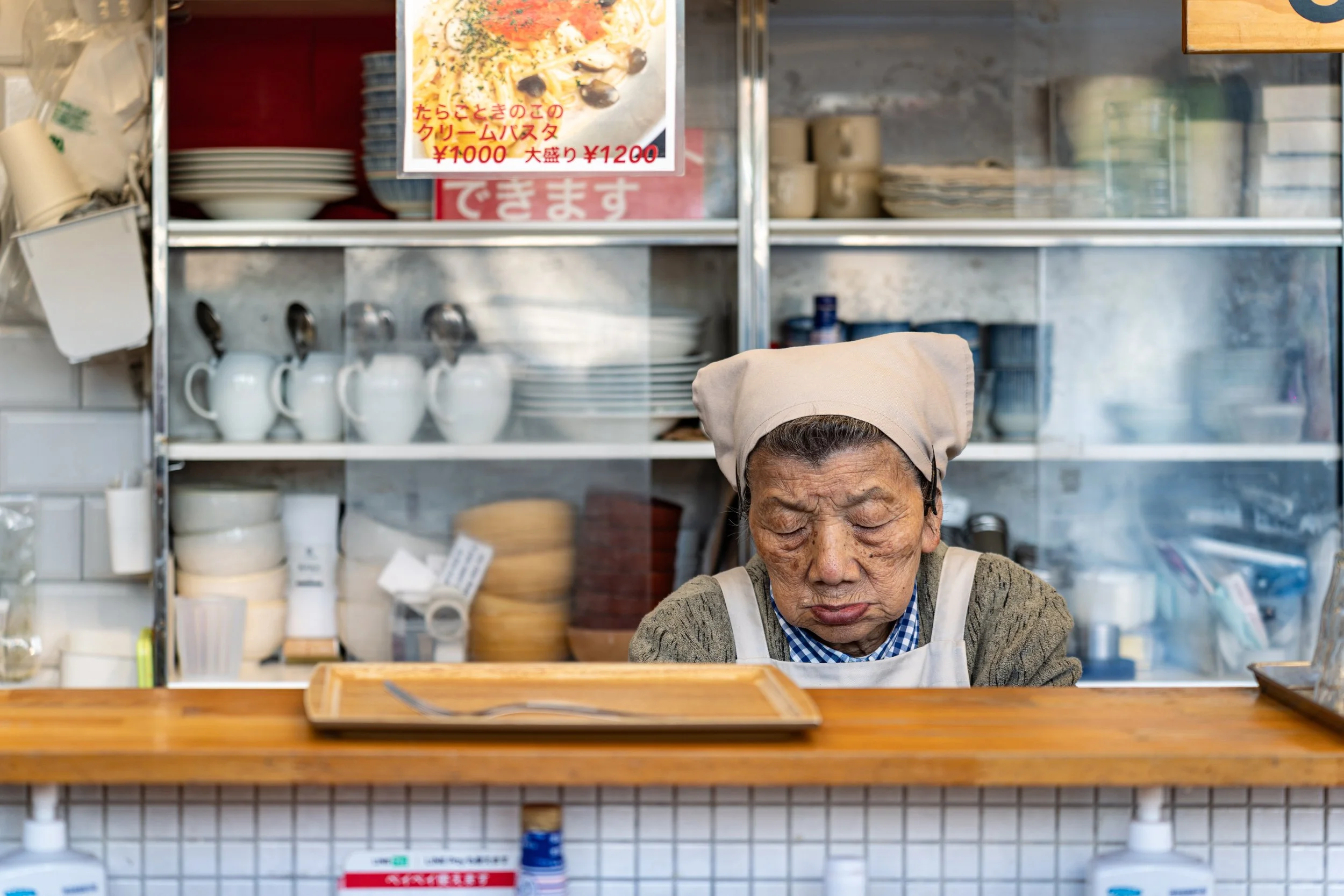 An elderly woman wearing a beige headscarf and apron, with her eyes closed, sitting behind a wooden counter in a kitchen or restaurant setting.
