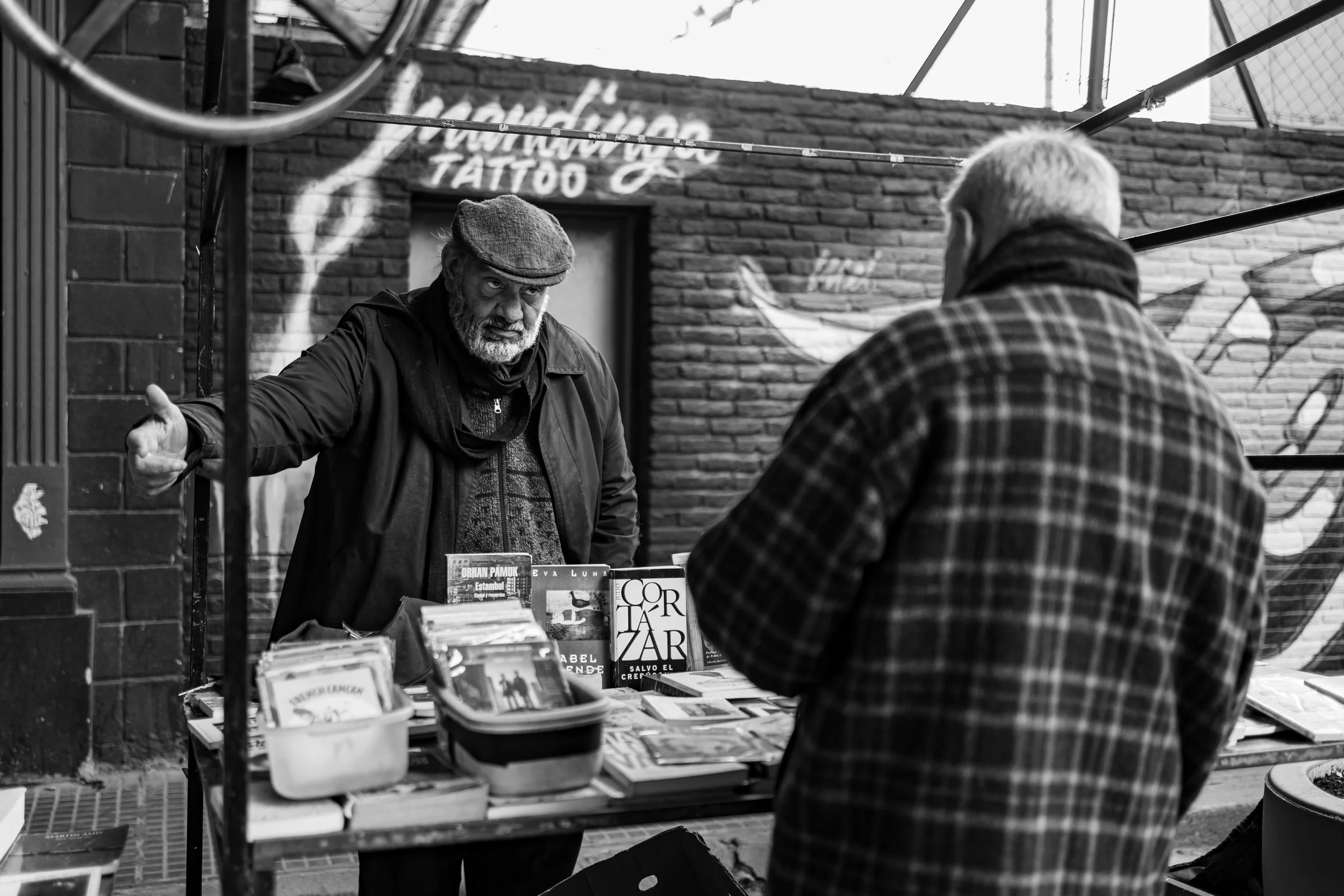 A street vendor with a beard and wearing a flat cap is conversing with a customer at a book stall on a sidewalk. The vendor is gesturing with his left hand while the customer, seen from behind, is wearing a checkered coat. Books are displayed on the stall, and a brick wall with a mural and signs is in the background.
