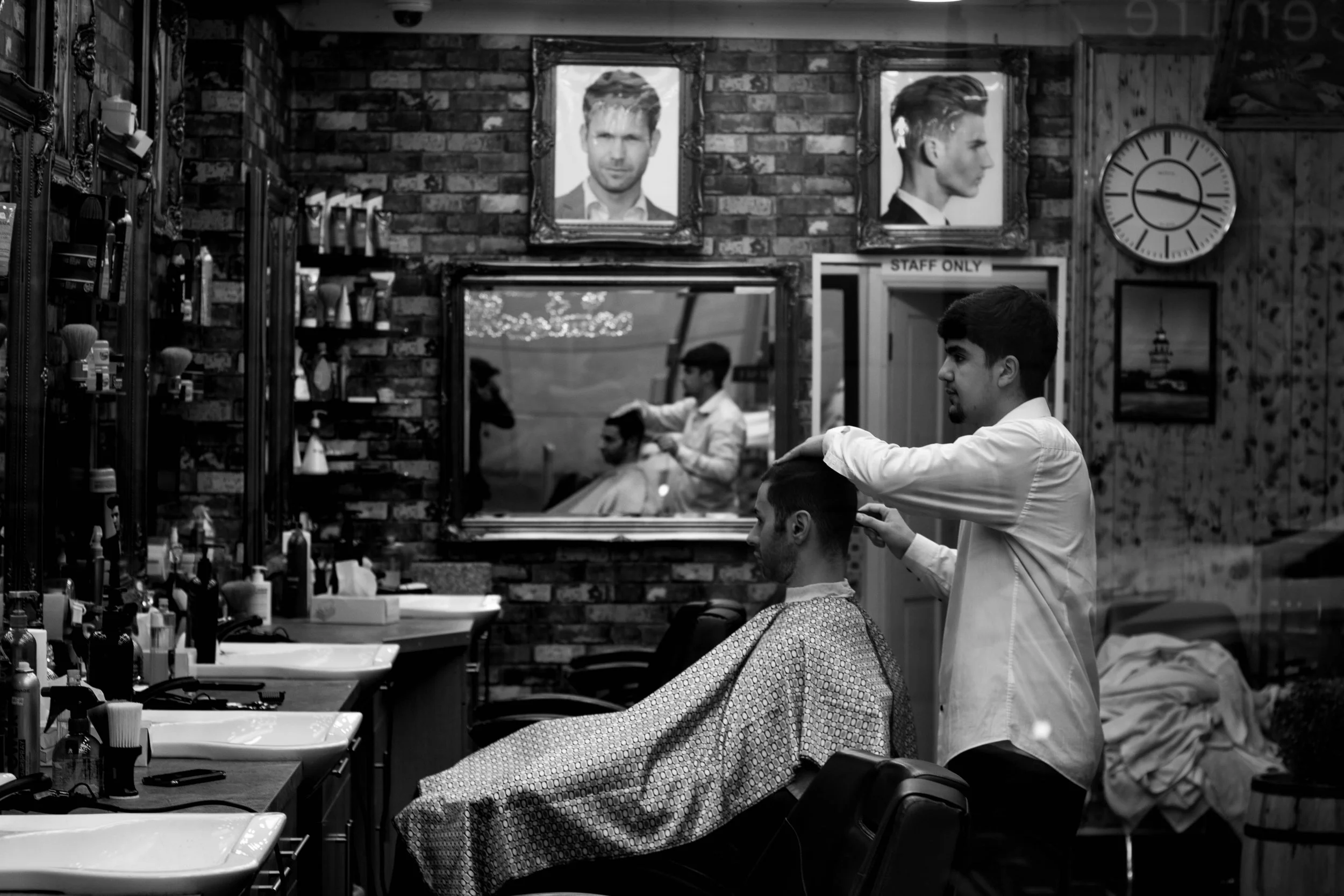 A man getting a haircut in a barbershop with brick walls, framed pictures, a mirror, and various hairdressing tools and supplies.
