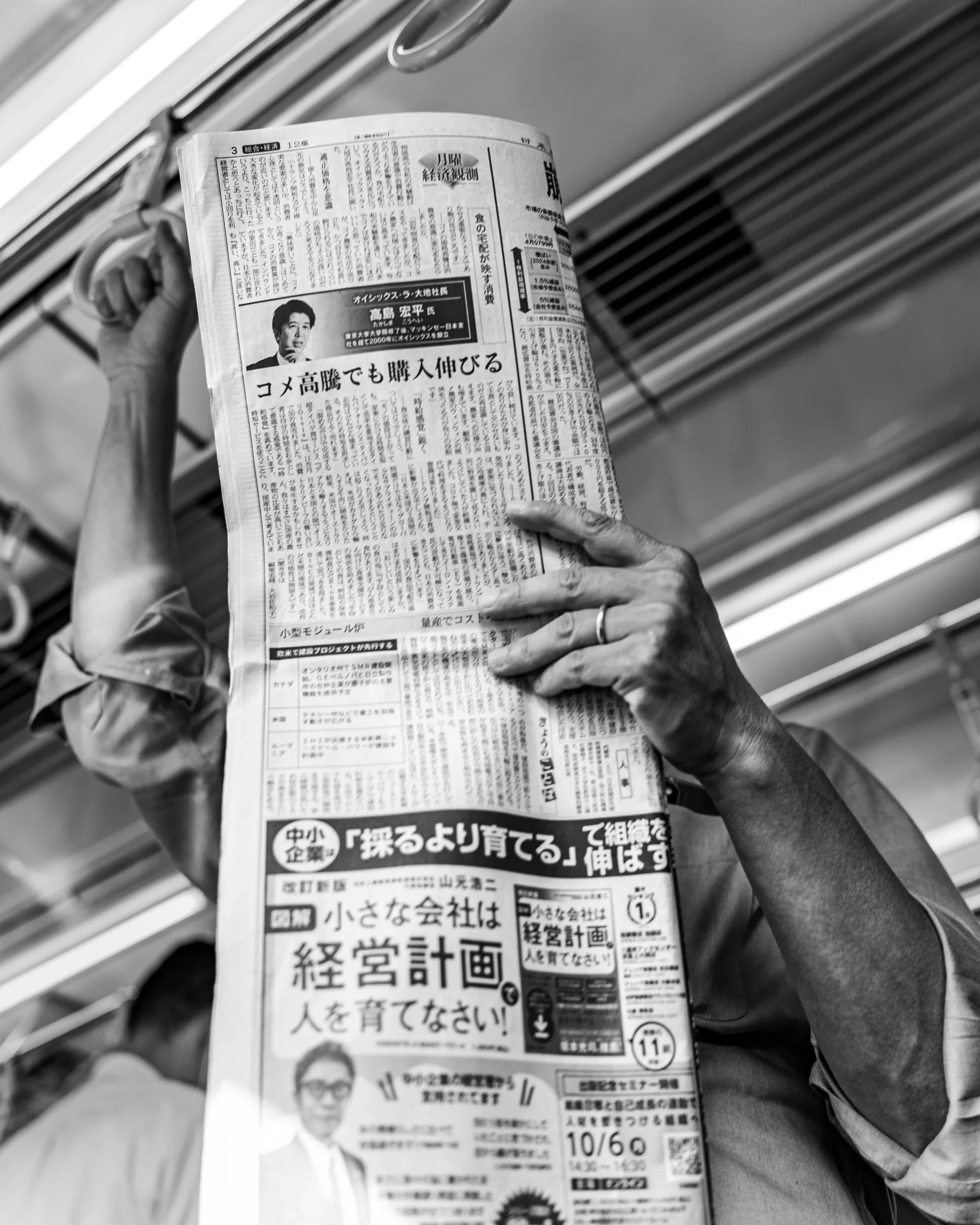 Person holding a Japanese newspaper while riding on a train or bus, with metal bars and ceiling vents visible in the background.