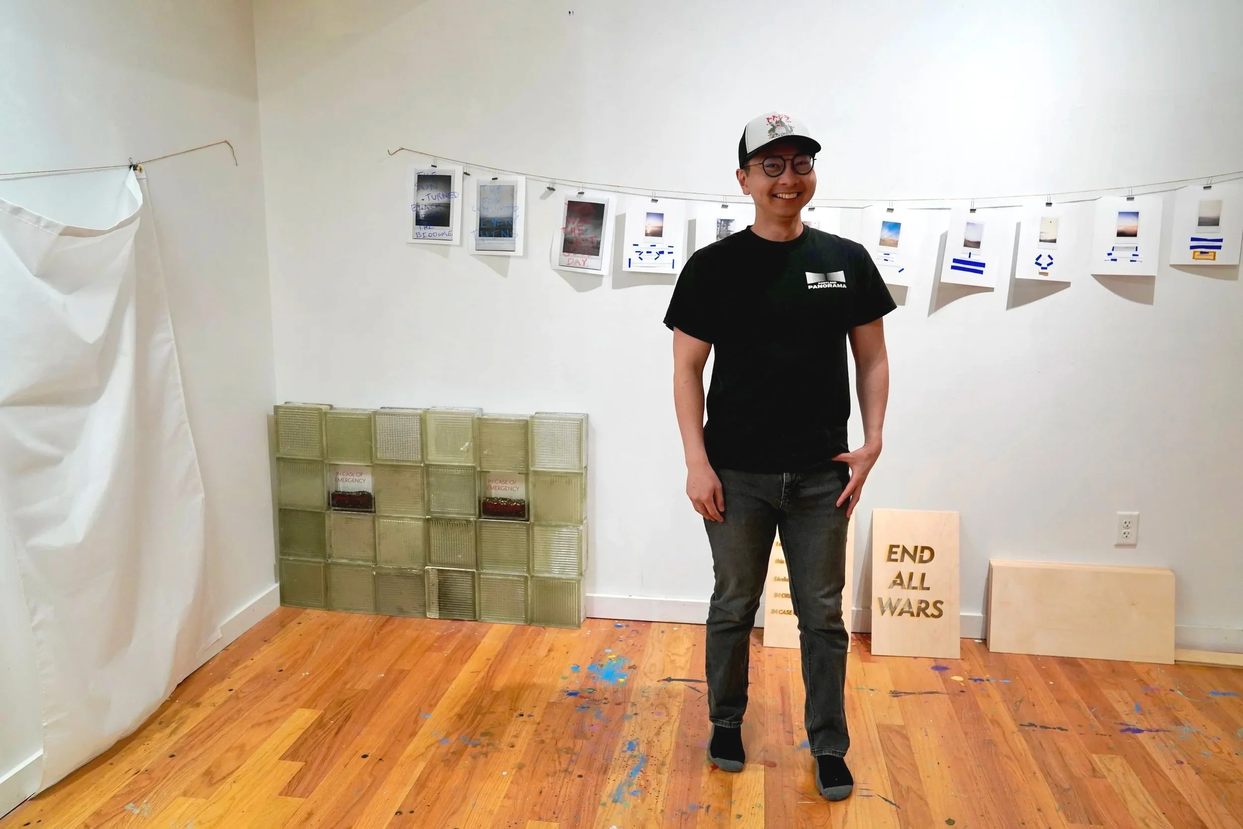 Taka standing in front of his work at stove works. stacked glass blocks in the corner. prints hang from strings along the back wall. and a wood panel has end all wars written on it.