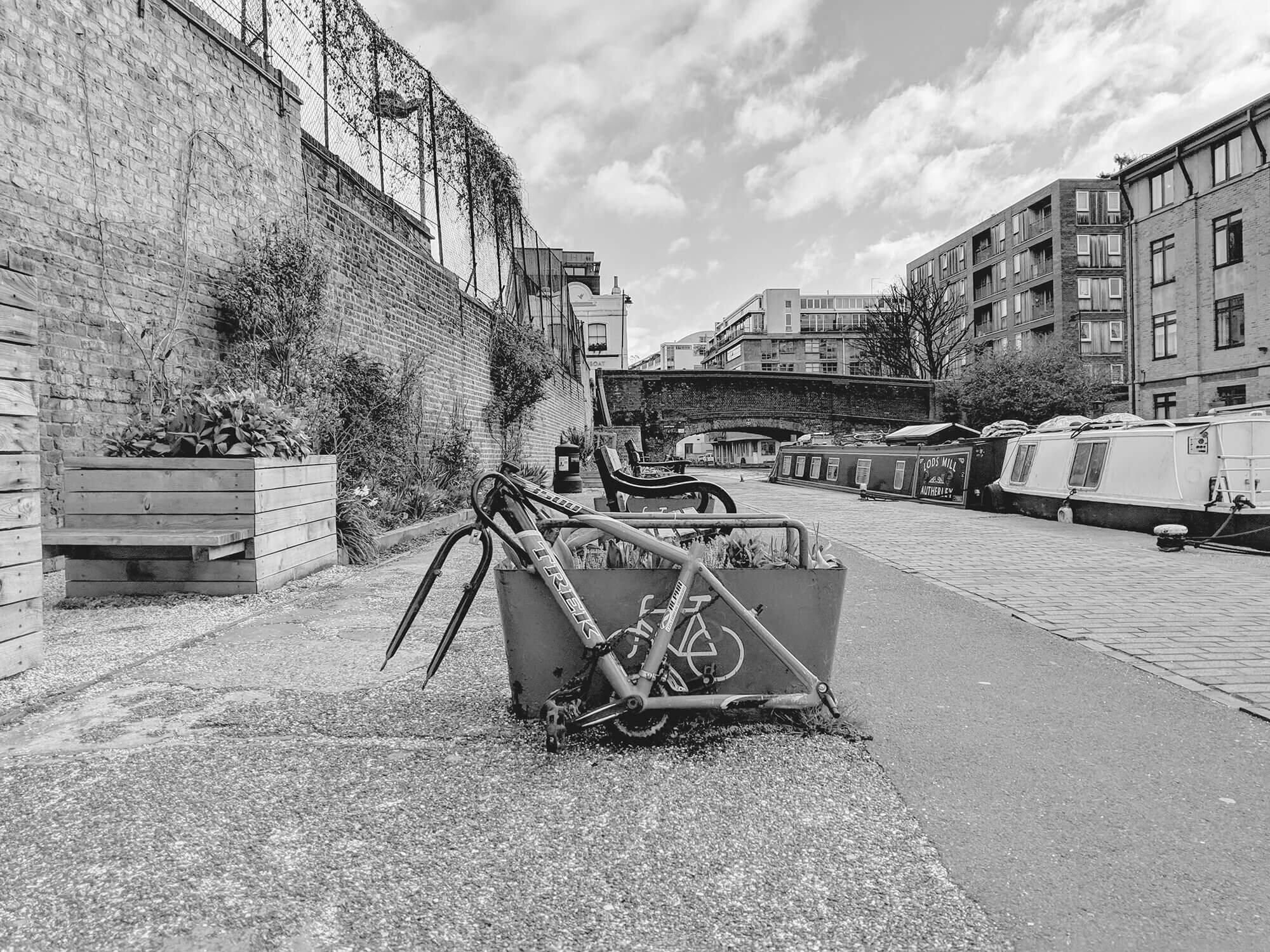 Bicycle with no wheels attached to flower box stand. Left abandoned on Regents Canal London.