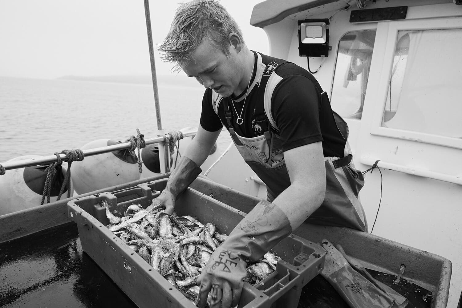 black and white photo of fisherman wearing silver necklace, sorting fish on a boat.