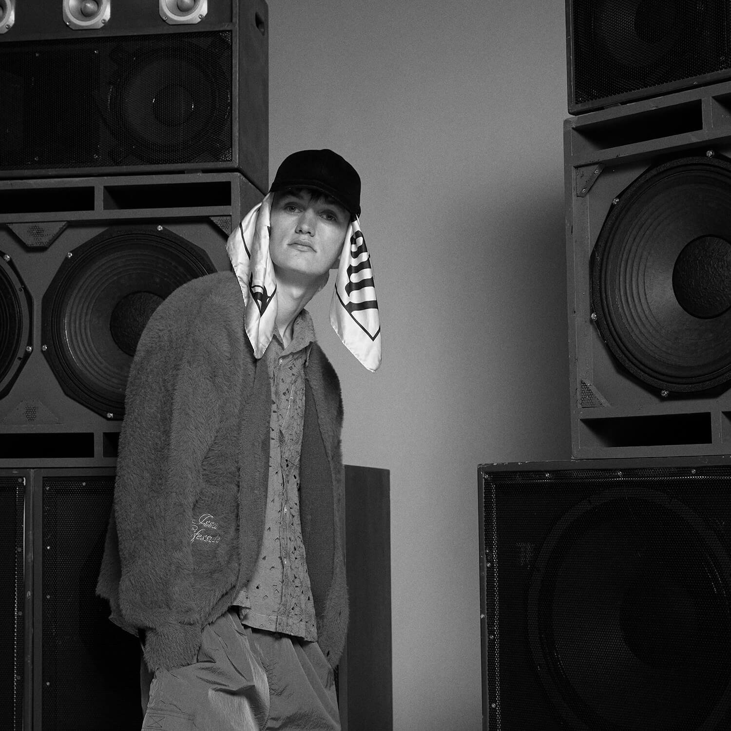 black and white photo of male model wearing cap and cardigan in front of a set of speakers.