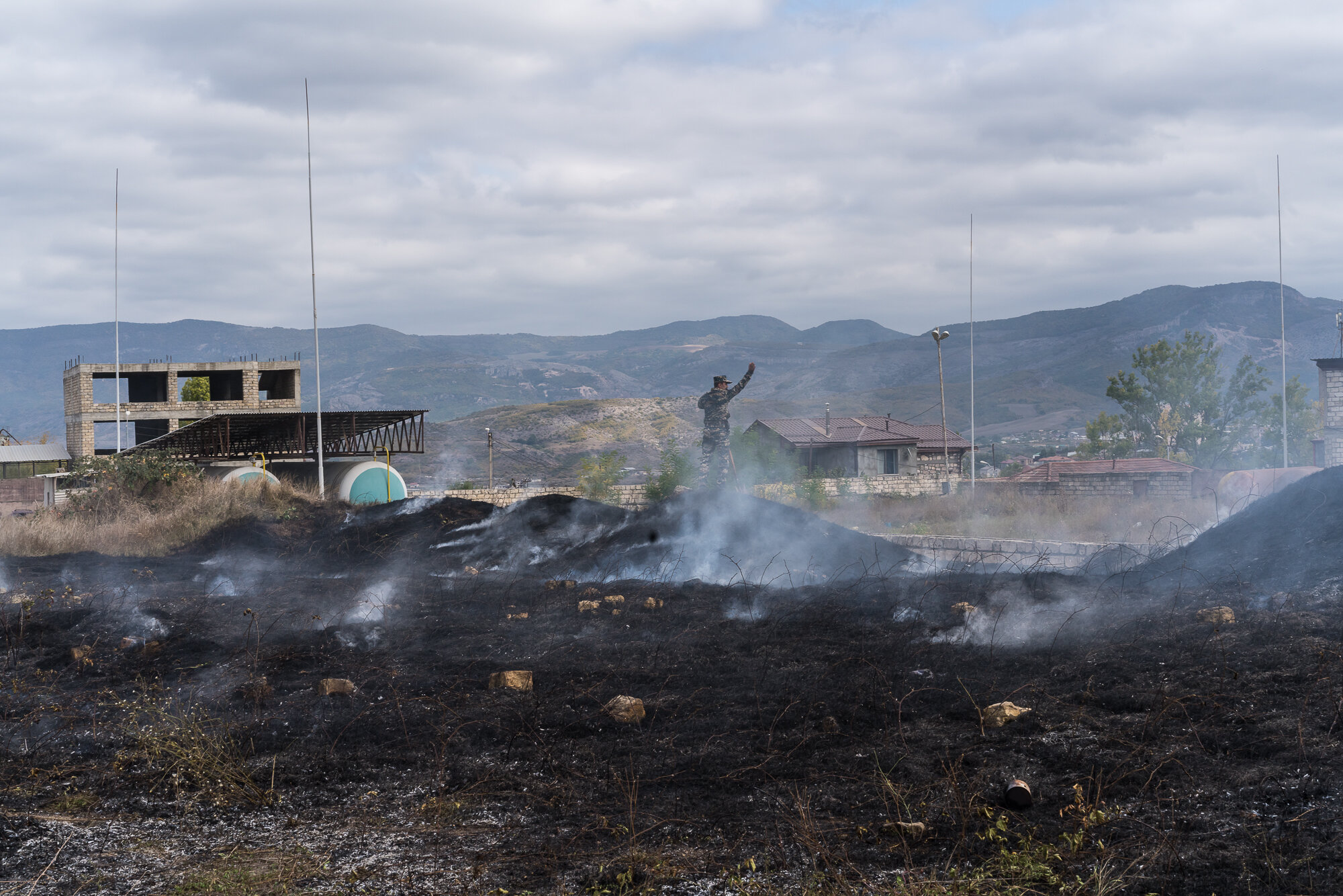  An emergency services member stands atop the site of a grass fire started by a bomb that fell nearby. Stepanakert, Nagorno-Karabakh. 2020.  