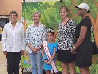 Group of five women and a young girl standing indoors in front of a green nature-themed backdrop, smiling at the camera.