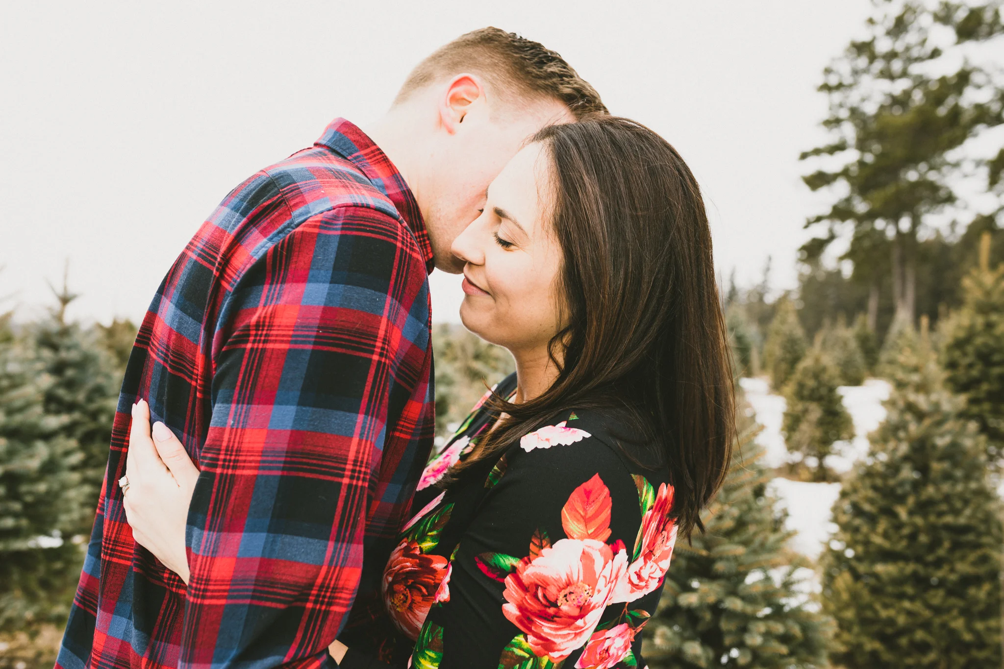 woman in black dress with red flowers hugging her husband at a tree farm - AMG Phototgraphy