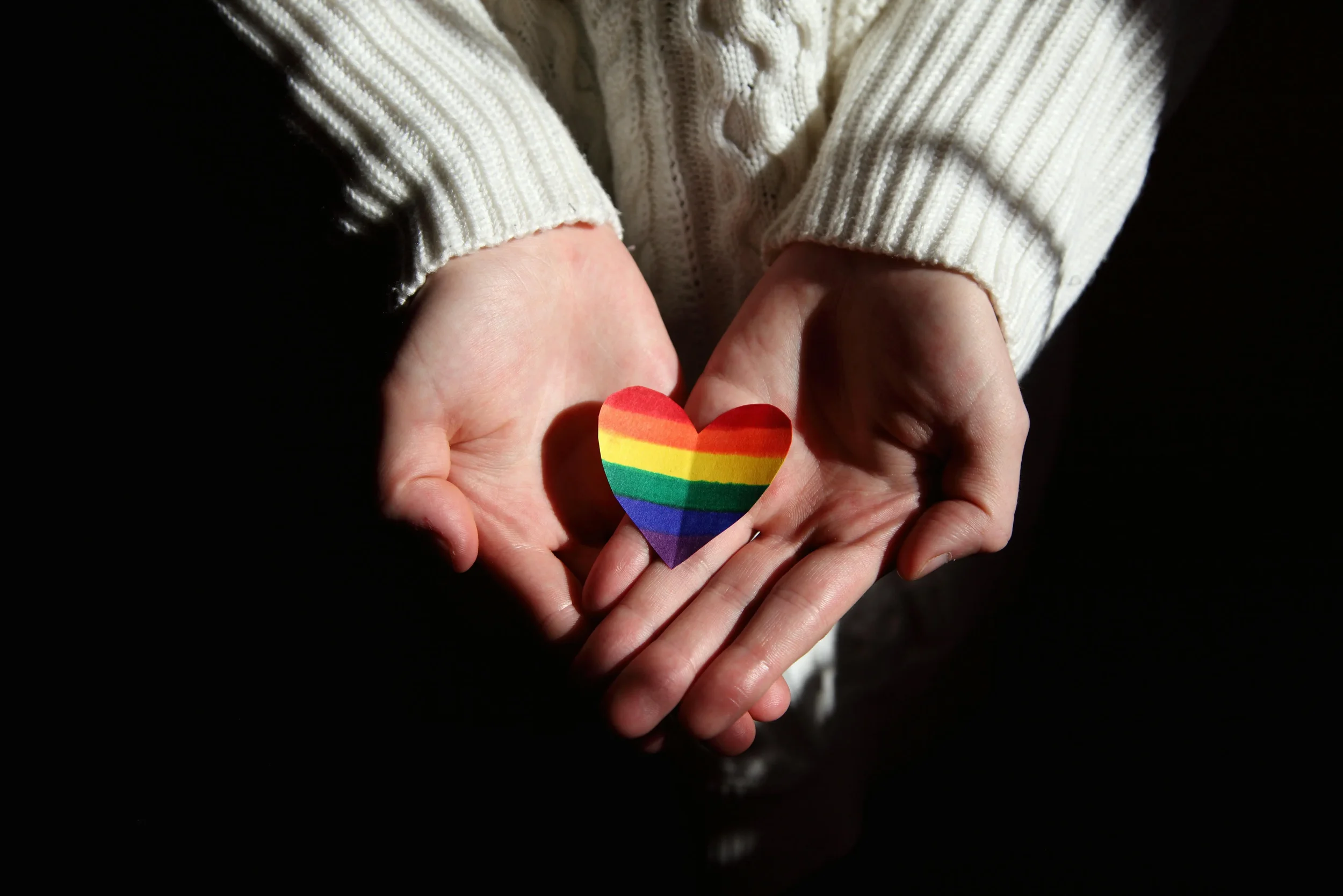 A hand holding a rainbow flag heart