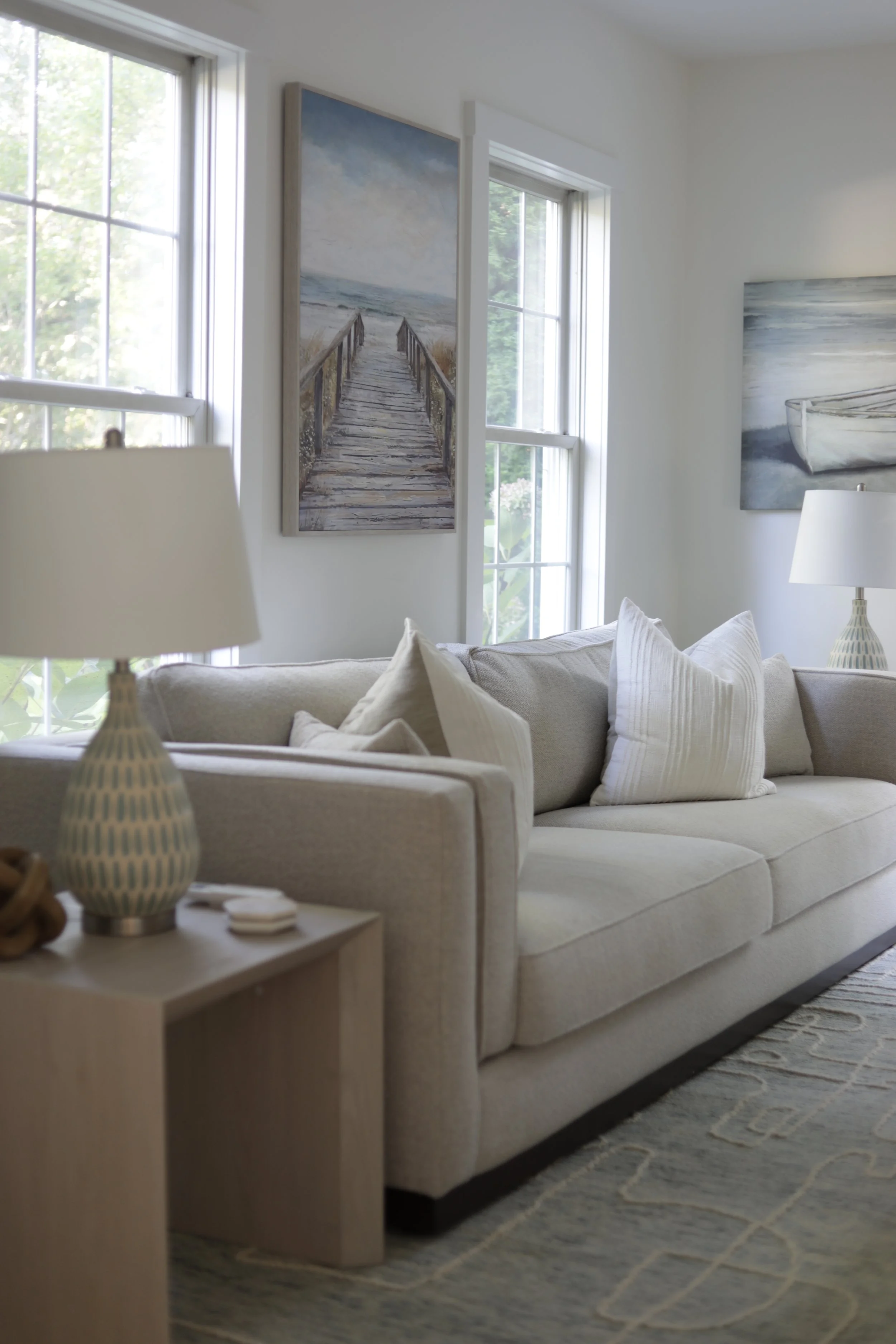 A living room with a beige sofa, white pillows, a side table with a lamp, and artwork on the wall featuring beach scenes, with large windows letting in natural light.