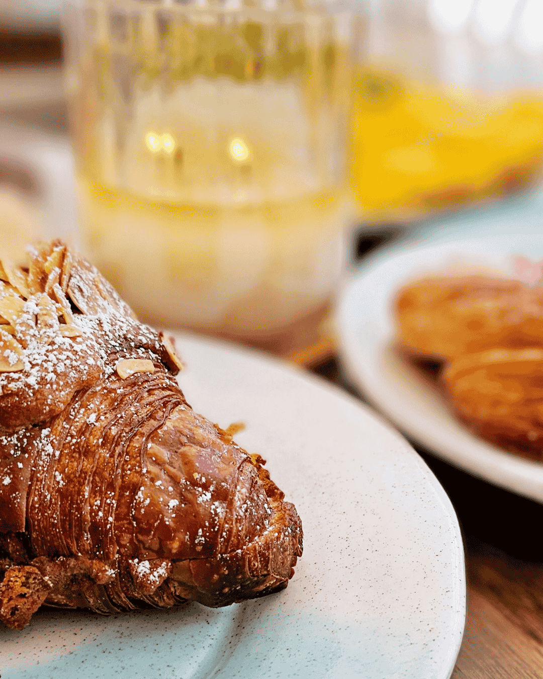 Close-up of a flaky, powdered Danish pastry on a white plate with a blurred background showing a glass of milk and other pastries.