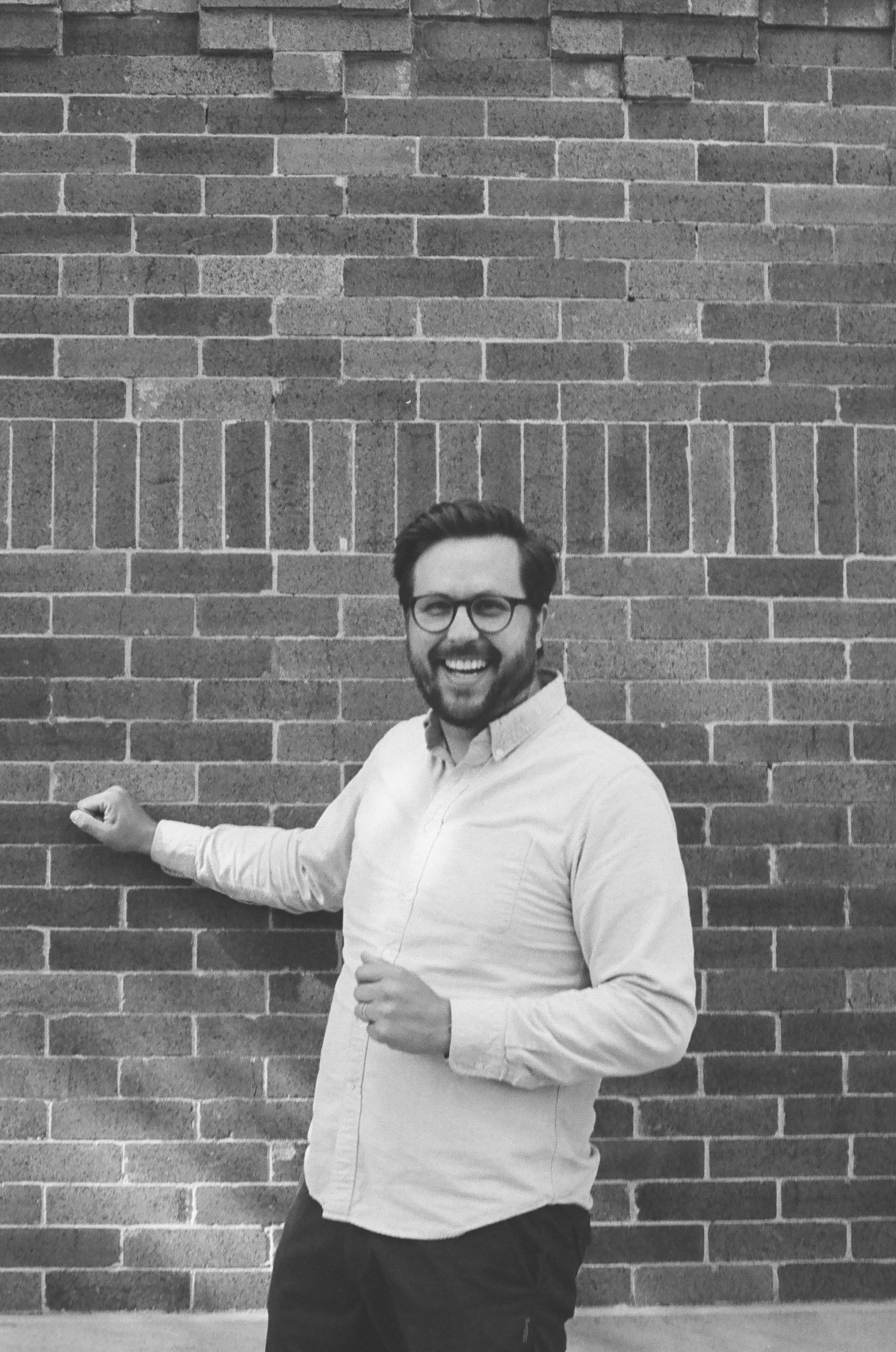 A photograph of a man standing in front of a brick wall smiling widely