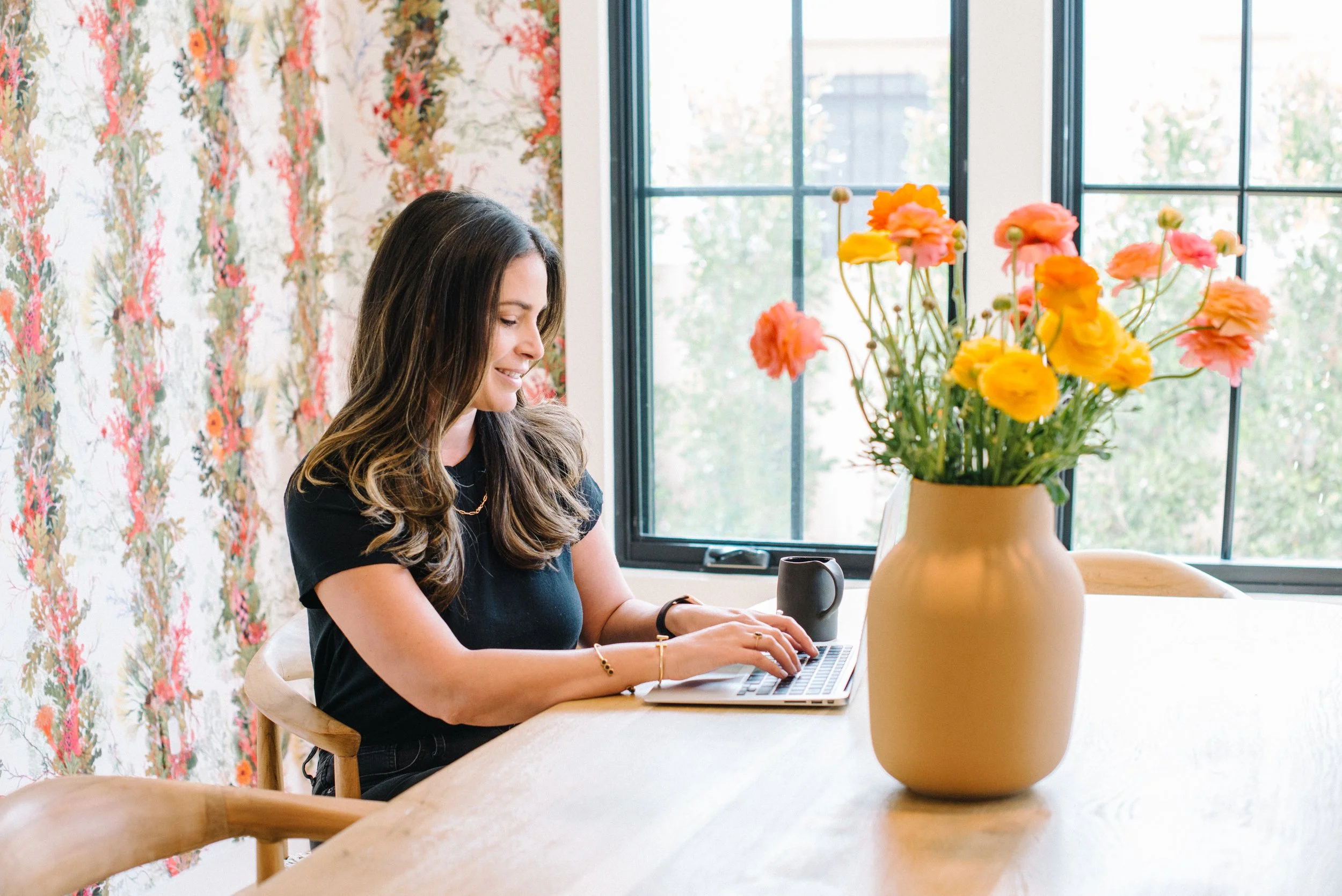 women with dark hair sitting at dining table with laptop and vase of fresh flowers
