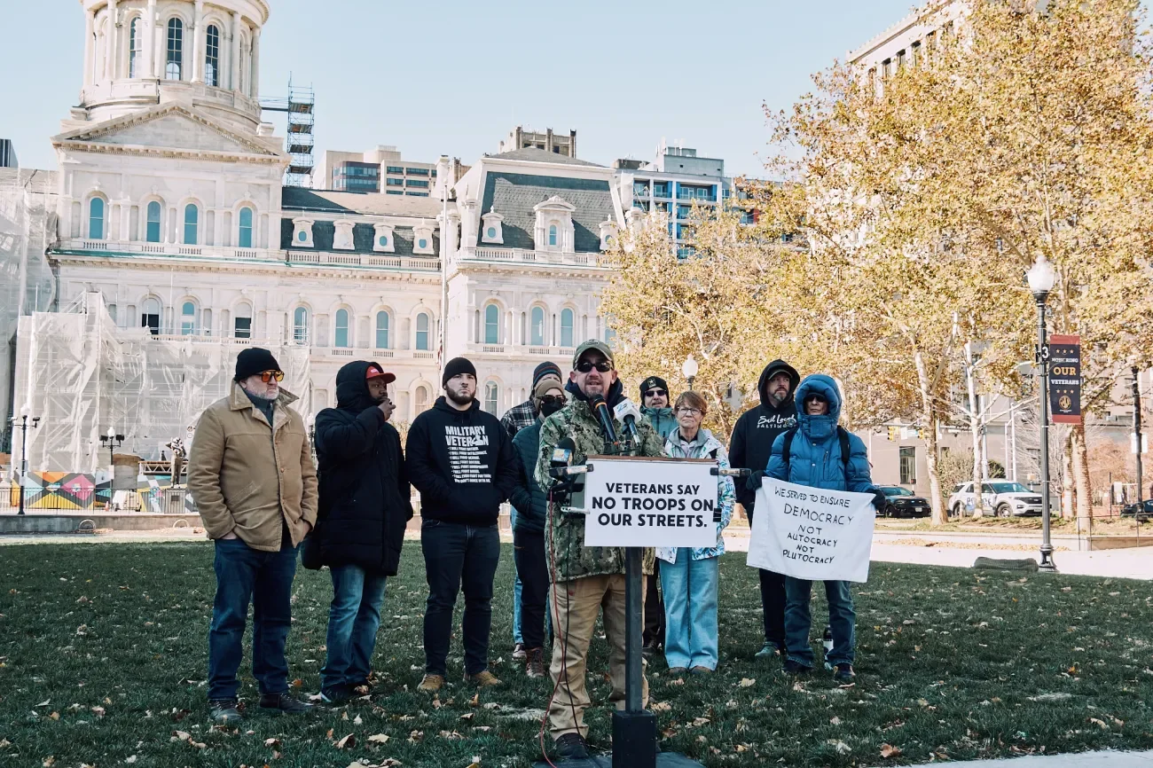 A group of activists speaking in front of a government building. A sign reads, "Veterans say no troops on our streets."