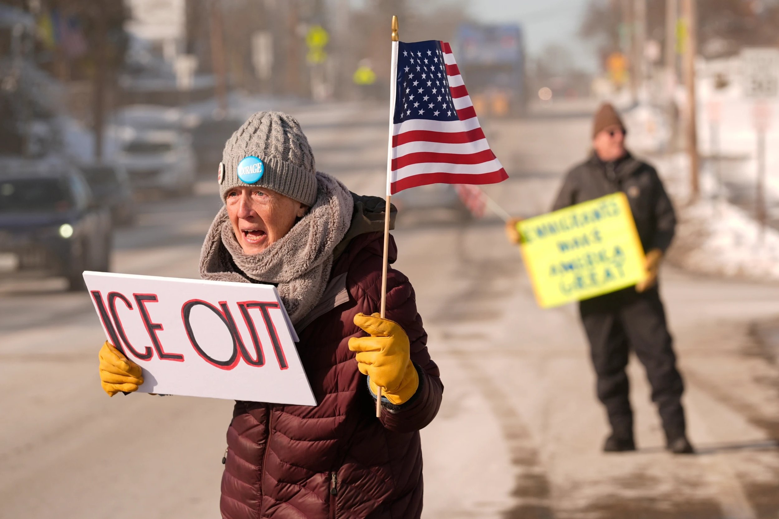 an older white woman stands in a snowy street with a US flag and a sign saying "ICE OUT"