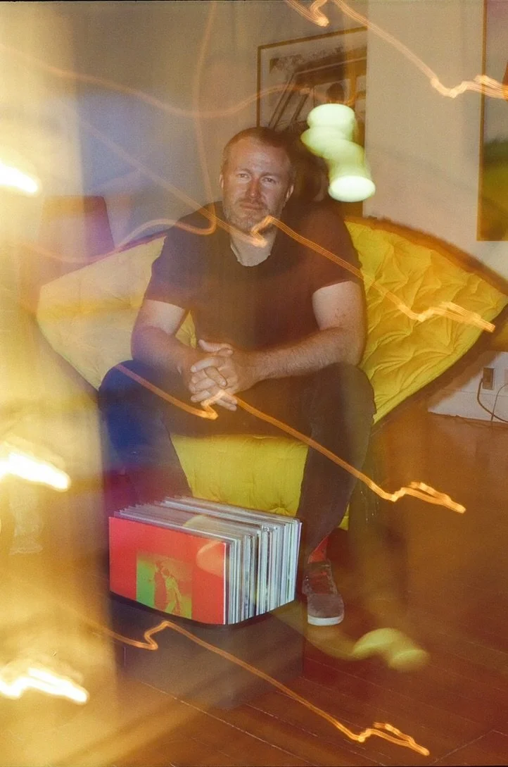 Man sitting on a yellow chair with a blurred background and streaks of light, next to a box of vinyl records on the floor.