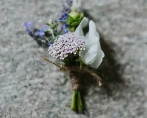 Close-up of a small, delicate summer wedding boutonniere wrapped in rustic twine, featuring a pale pink yarrow or lace flower, a single white bloom, and tiny blue wildflowers on a textured grey background.