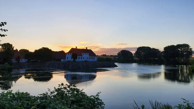 A landscape view of the historic Soebygaard manor house surrounded by its calm castle moat under a sunset sky. A scenic cultural attraction for couples on an elopement honeymoon on Aeroe Island in Denmark.