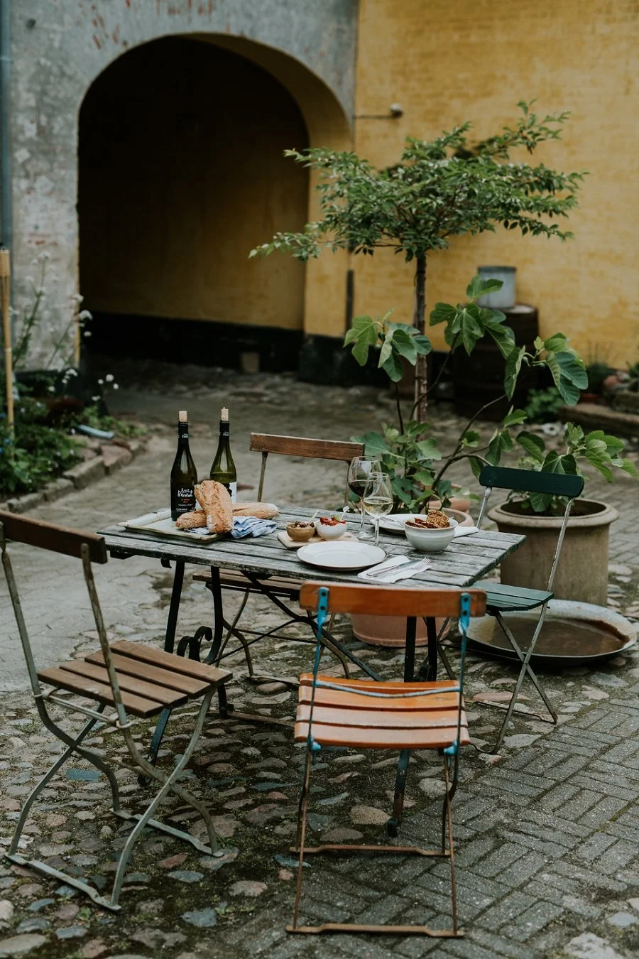 A cozy outdoor table setting in the historic courtyard garden at Den Gamle Kobmandsgaard in Aeroeskoebing. Featuring fresh bread, wine glasses, and local tapas treats for couples enjoying their elopement honeymoon on Aeroe Island in Denmark.