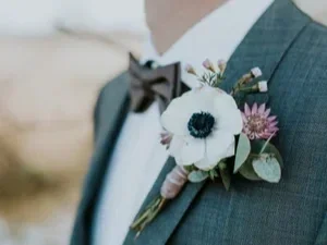 Close-up profile of a smiling groom wearing a grey textured suit and a brown bowtie, featuring an elegant spring wedding boutonniere with a white poppy anemone, a purple wildflower, and eucalyptus leaves.