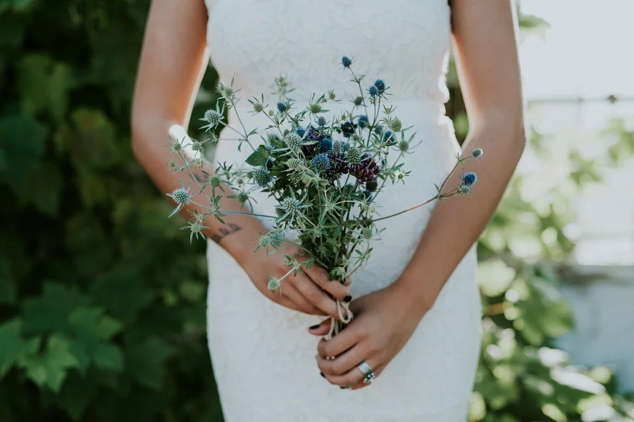 A close-up shot of a bride in a white wedding dress holding a delicate bouquet of local wildflowers and blue thistles.
