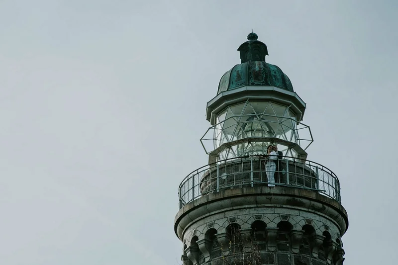 A wedding couple standing on the top gallery of Skjoldnaes Fyr lighthouse. A dramatic, wild coastal location for an outdoor destination wedding ceremony or sunset honeymoon view on Aeroe Island in Denmark.