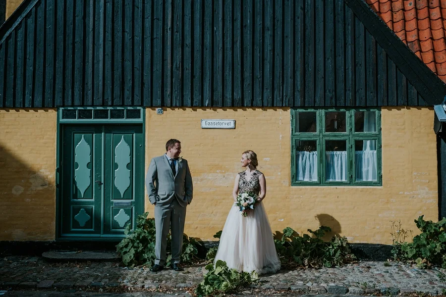 A wedding couple standing on the cobblestone street in front of a historic yellow half-timbered house with a green wooden door in Aeroeskoebing. An iconic town setting for an elopement wedding on Aeroe Island in Denmark.