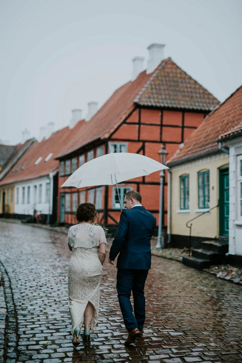 A wedding couple walking under a white umbrella on a rainy day in a rainy street of Aeroskobing.