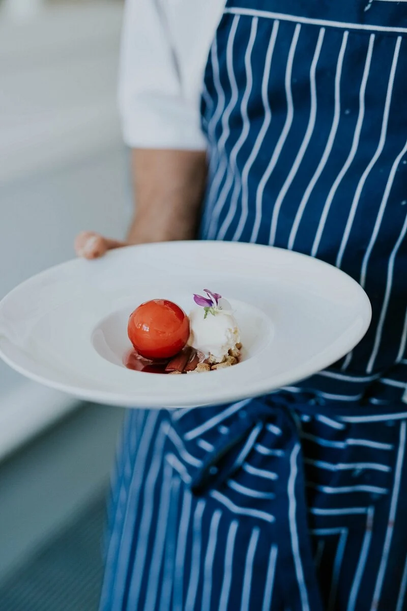 A beautifully presented gourmet dessert with a sorbet sphere and cream on a white plate, served by a chef in a striped apron. High-end fine dining inspiration for an elopement honeymoon on Aeroe Island in Denmark.