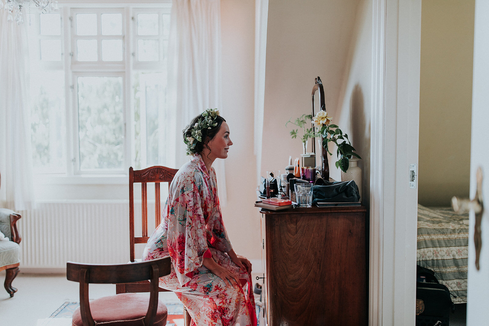 A bride wearing a floral crown and a patterned kimono robe sits by the vanity table while getting ready for her micro wedding in Denmark