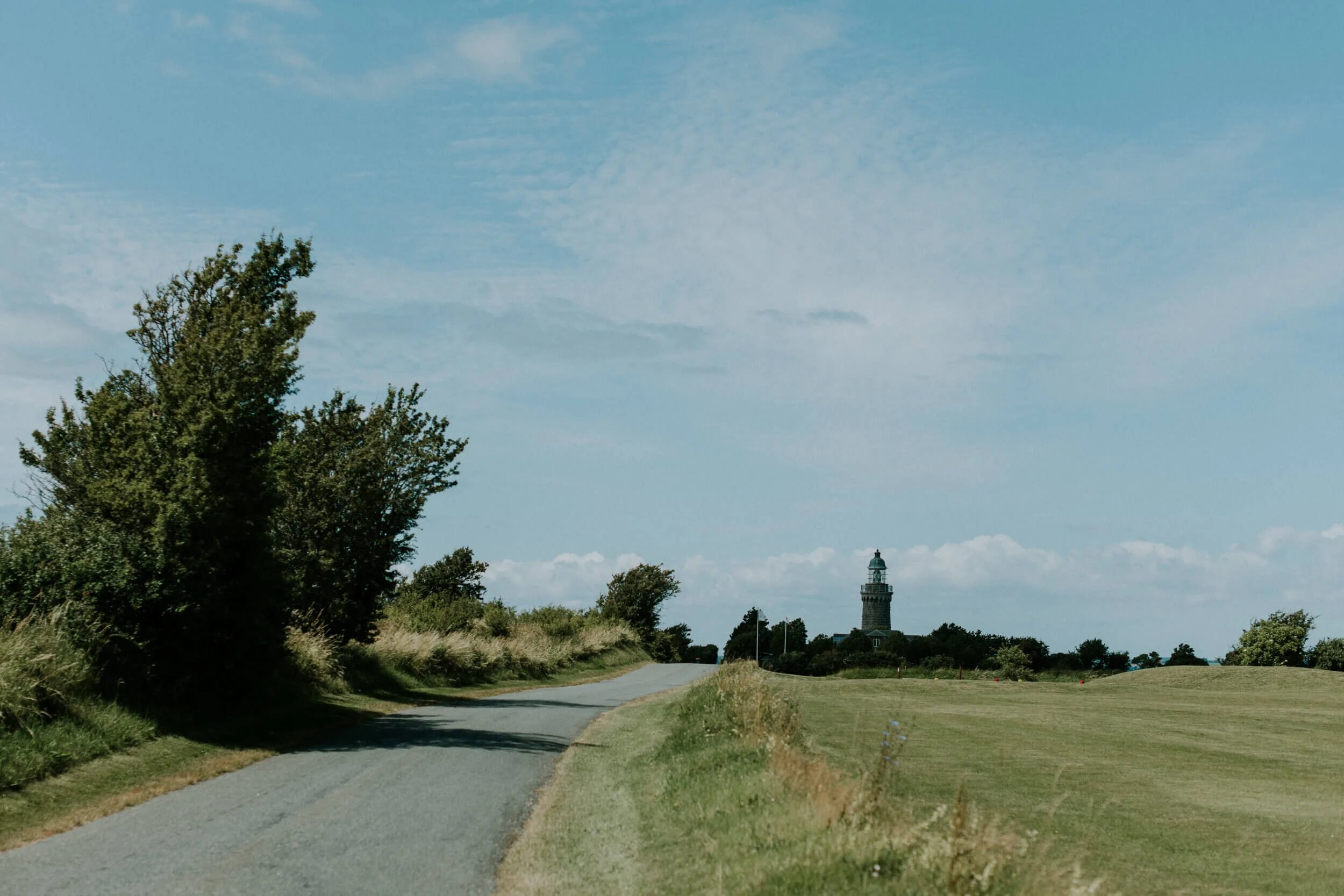A scenic view of the winding asphalt road leading towards Skjoldnaes Fyr lighthouse surrounded by green fields. A beautiful coastal landscape detail for couples exploring Aeroe Island during an elopement honeymoon in Denmark.