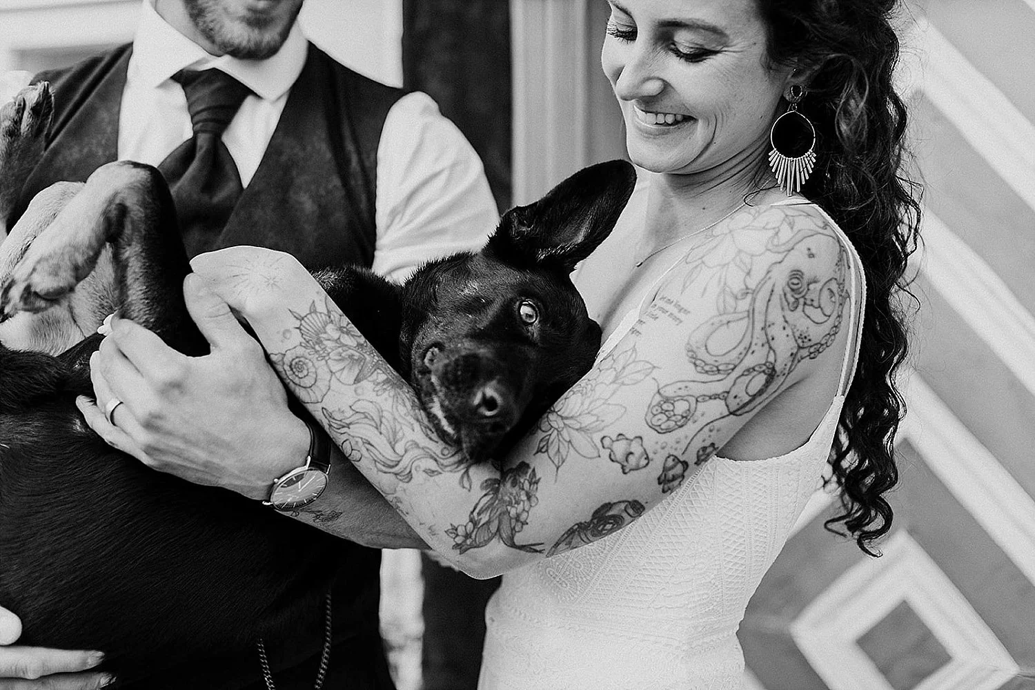 Black and white wedding photo of an international couple holding their happy black dog, leaning against the bride's chest.