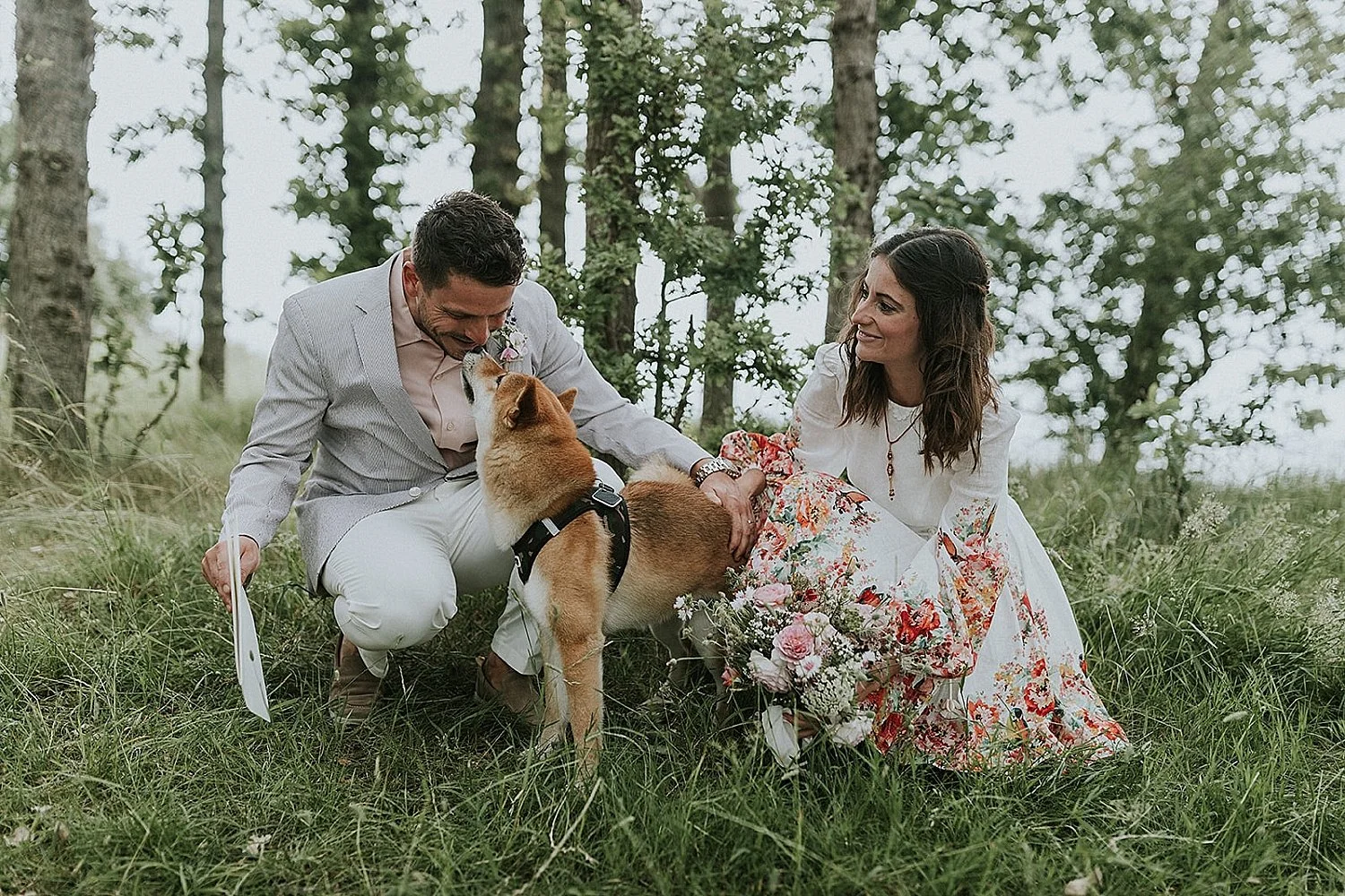 Newlyweds crouching in a small grove with their dog touching noses with the groom during an outdoor wedding in Denmark.