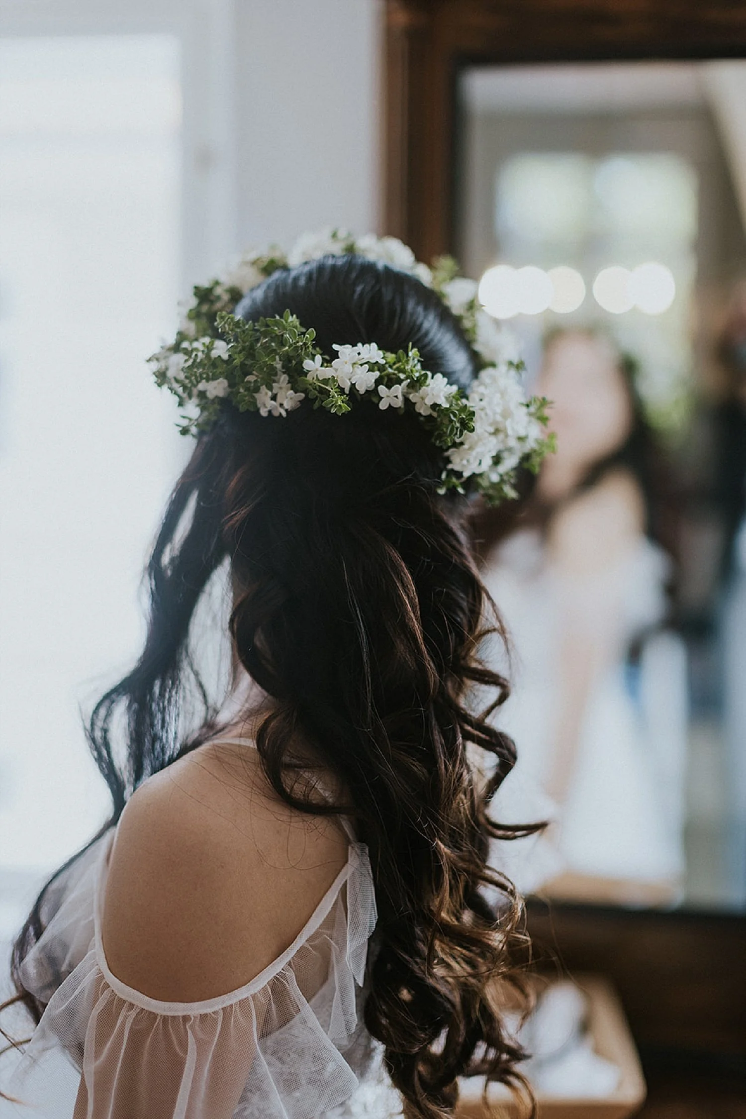 Bride from behind in the beauty room mirror with long bridal curls and a seasonal white flower crown