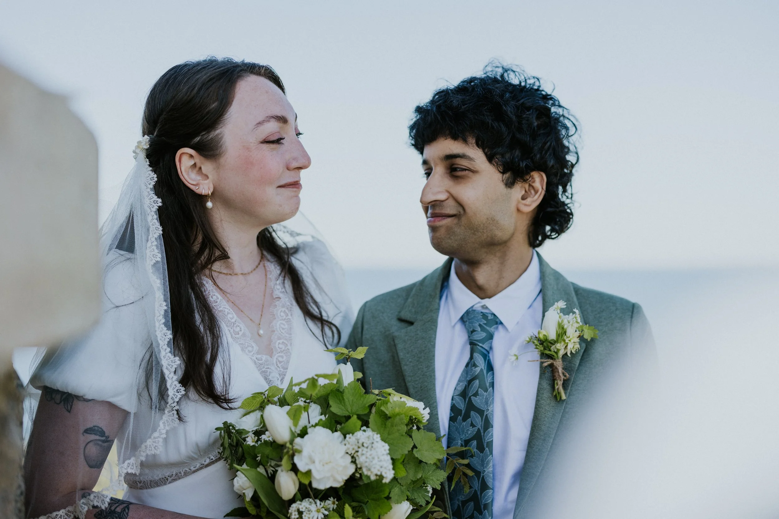 Smiling wedding couple looking at each other, highlighting the groom's green suit jacket and a simple white boutonniere with fresh green foliage.