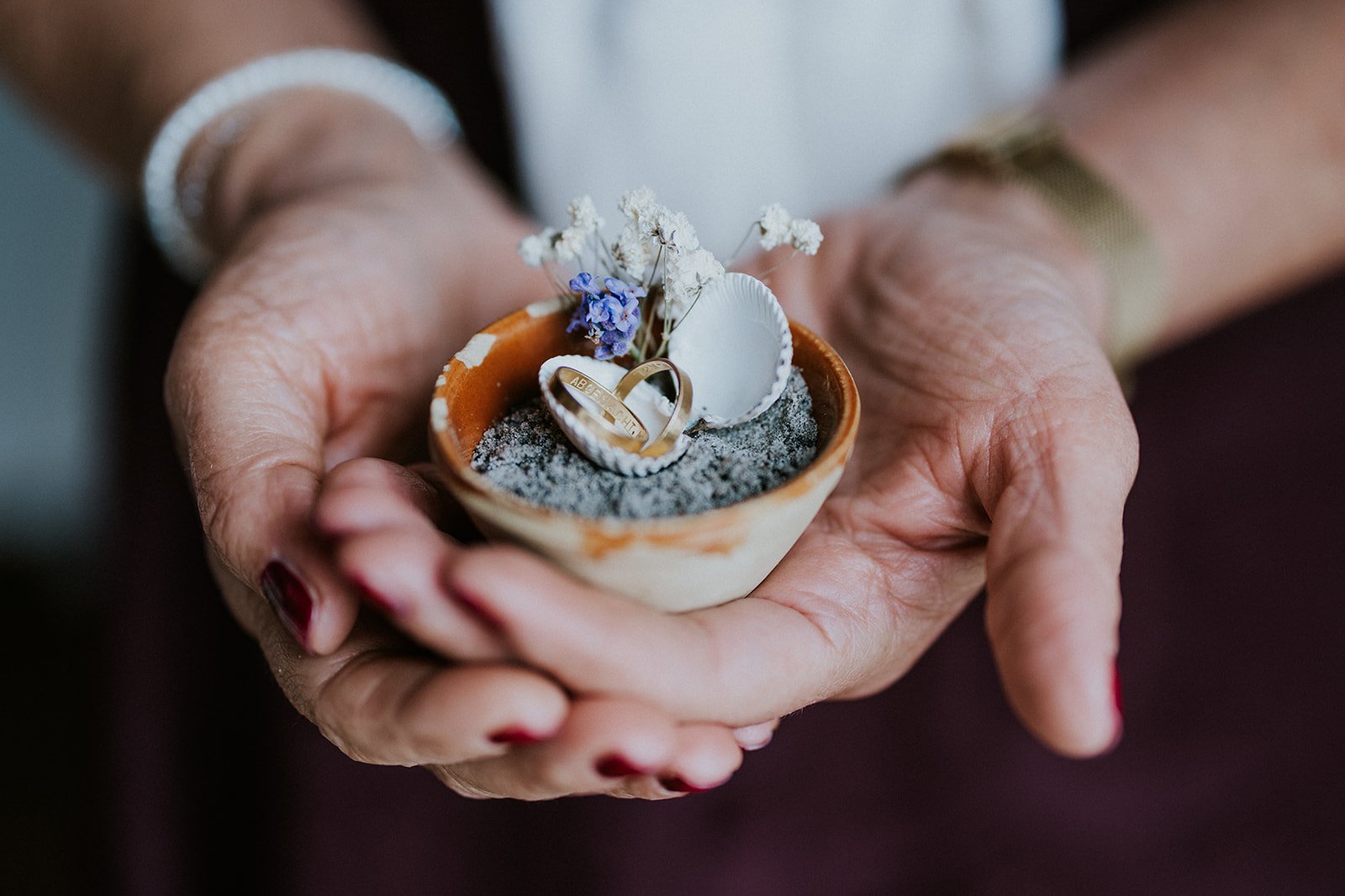 A close-up shot of wedding rings placed inside a small seashell with dried flowers held in hands.