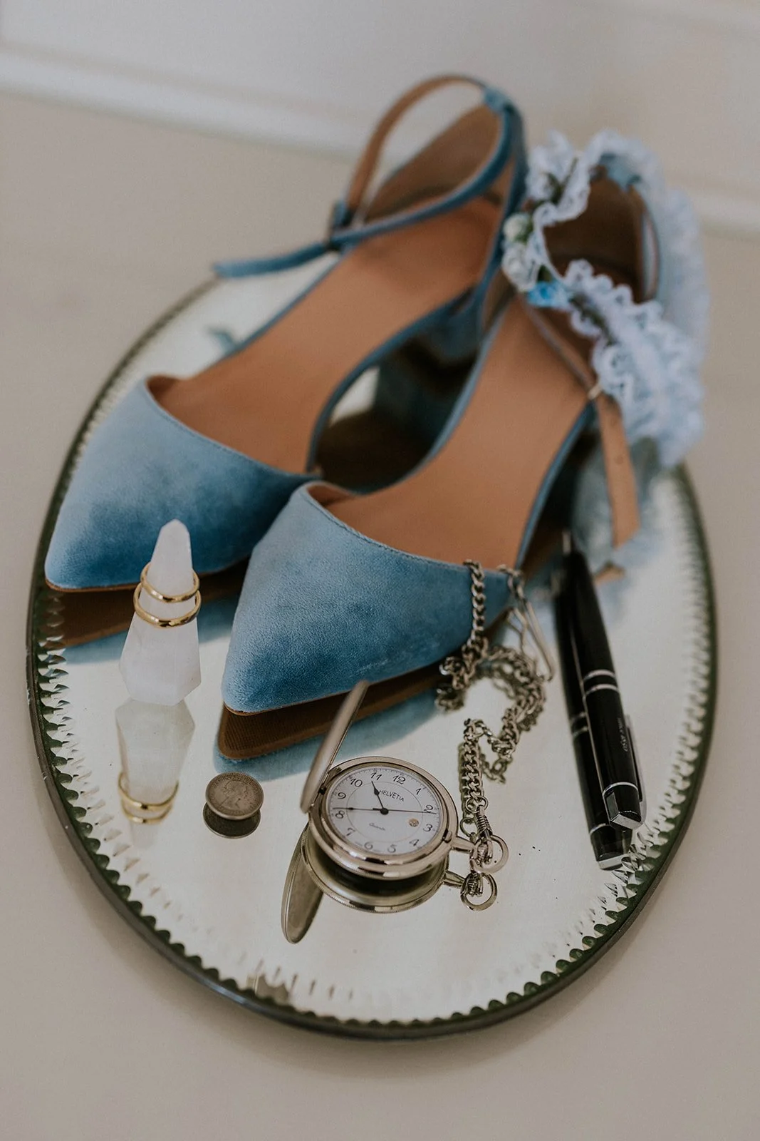 Blue bridal shoes placed on a vintage mirror alongside gold wedding rings, an antique pocket watch, a fountain pen, and a historic sixpence coin.