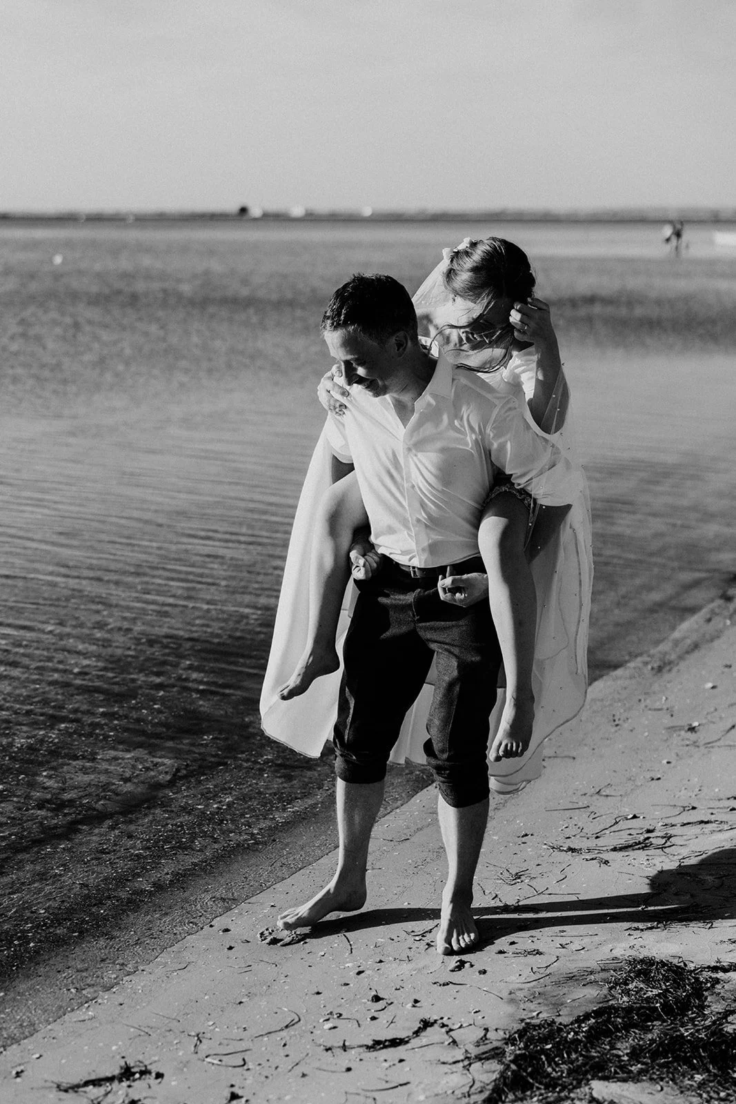 A black and white photo of a barefoot bride and groom in wedding attire at the water's edge at Vesterstrand beach in Aeroskobing on Aero, with the bride riding piggyback as they laugh by the calm sea.