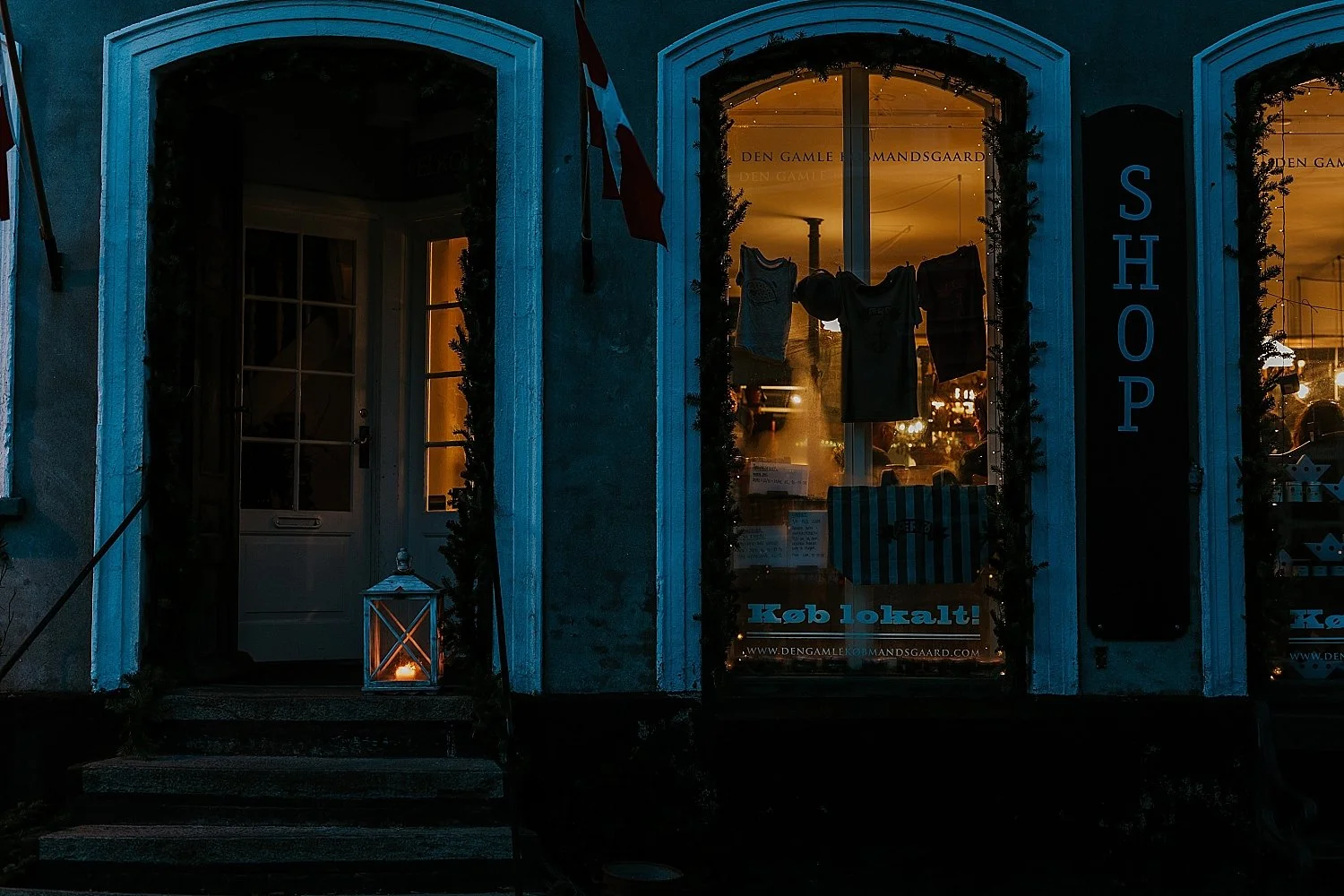 The illuminated storefront of Den Gamle Kobmandsgaard during a winter evening in Aeroeskoebing. Showcasing traditional Danish hygge, candles, and winter elopement honeymoon atmosphere on Aeroe Island in Denmark.