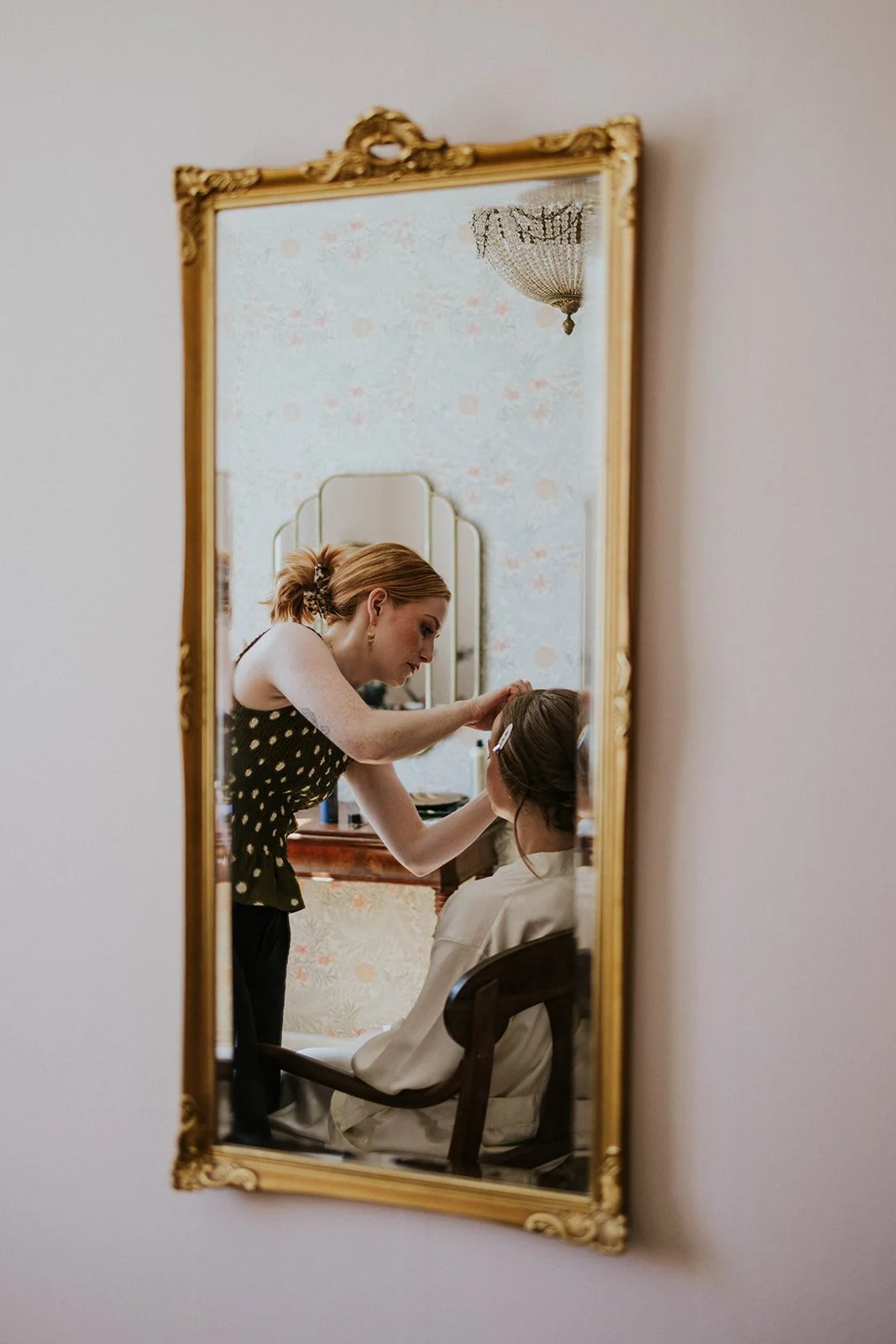 A reflection in an ornate gilded mirror showing redhead stylist Augusta applying wedding makeup and styling the bride in the beauty room.