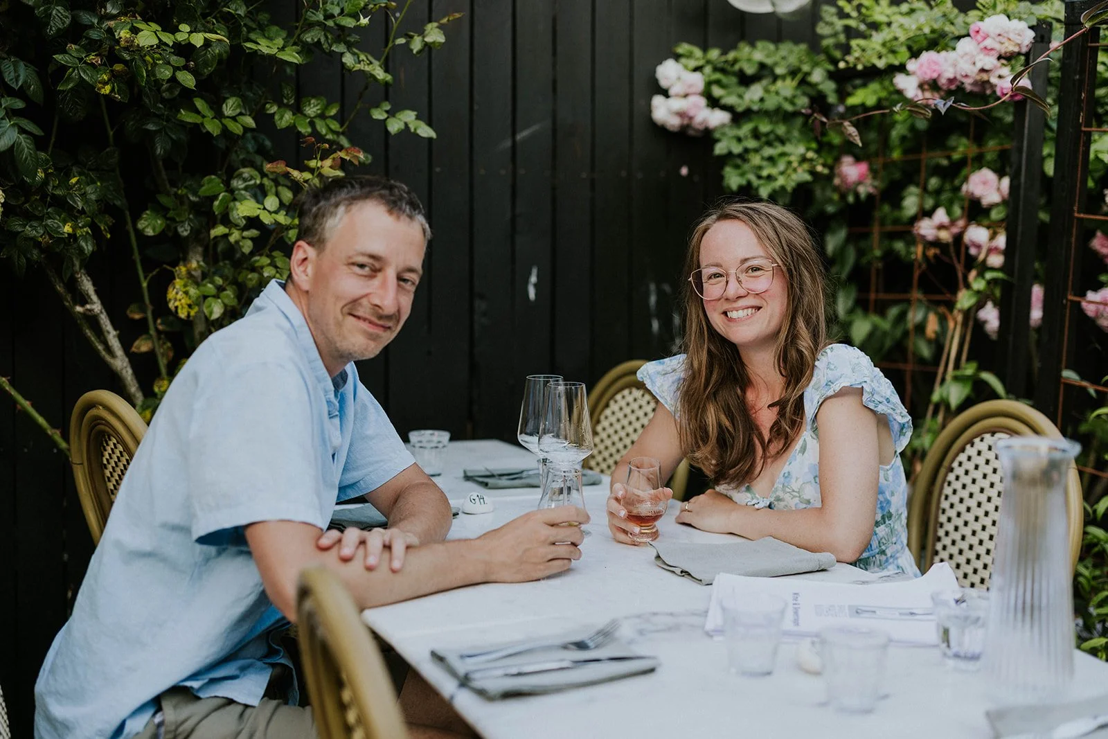 The bride and groom sitting at a table with a white tablecloth in the courtyard garden of Bangs Mad og Vinbar in Aeroskobing, enjoying an aperitif against a background of black wood and pink roses.