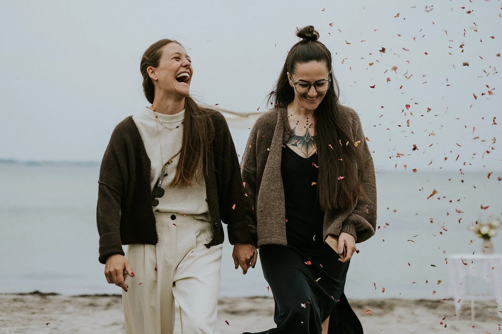 Two brides laughing and holding hands during a lesbian beach wedding on Aero Island in Denmark, with falling red rose petals