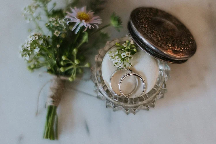 Close-up of a minimalist summer wedding boutonniere next to a silver ring box with two silver wedding rings on a white silk background.
