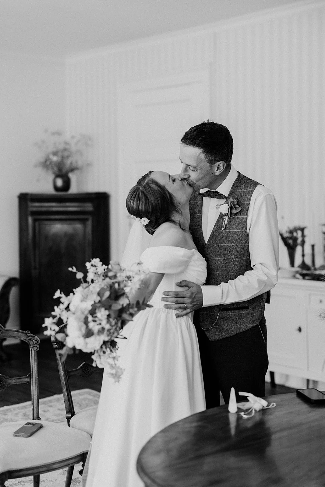 A black and white photo of the bride and groom kissing after their ceremony in the wedding hall at The Old Merchant's Court, surrounded by empire style furniture, oriental rugs, and the bride holding a large bouquet of local seasonal flowers.