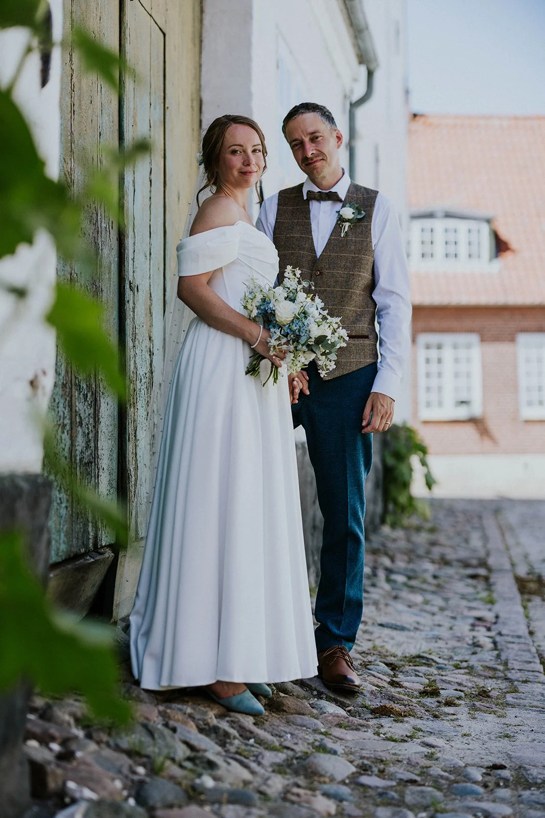 The bride and groom standing in a cobblestone street in Aeroskobing against an old door with peeling green paint, with hollyhocks in the foreground and a matching local flower bouquet and boutonniere.